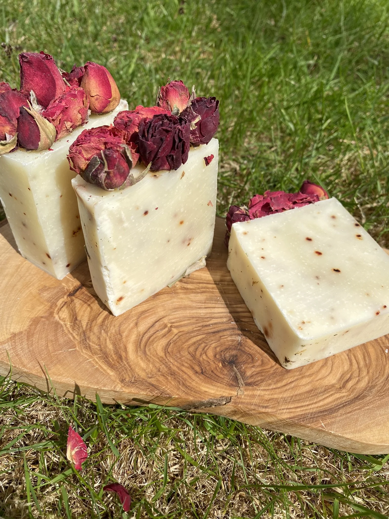 Bars of handmade soap with dried rosebuds on top, placed on a round wooden surface outdoors on grass.