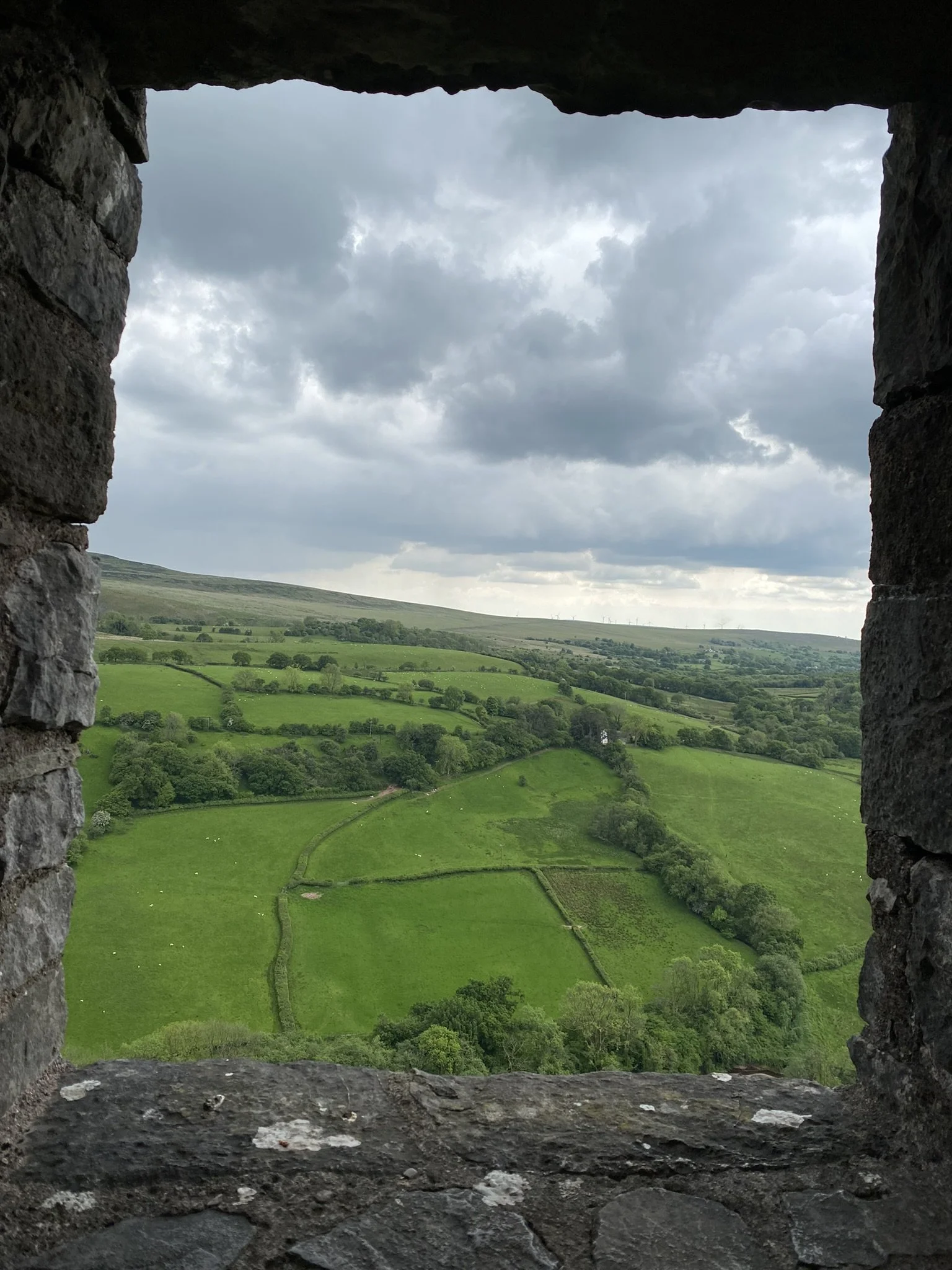 A view of lush green rolling hills and fields through a rectangular gap in a stone wall. The sky is overcast with dark clouds.