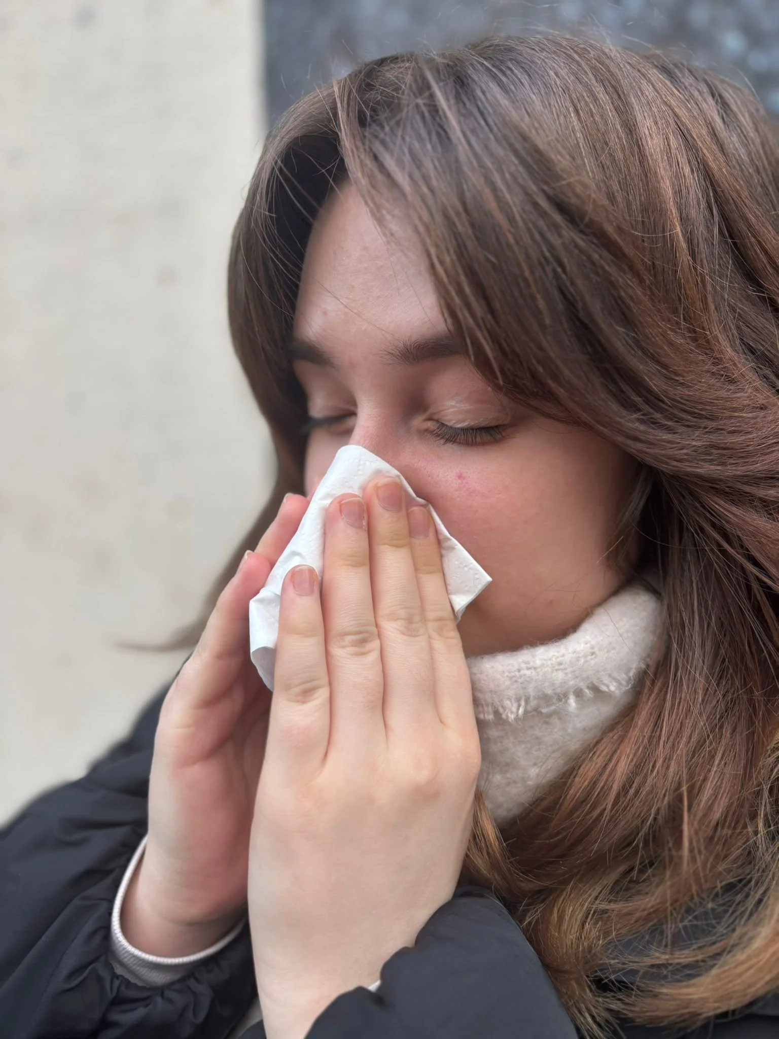 A person with long brown hair holding a tissue near their nose, possibly wiping or blowing their nose. Another person with light skin is gently supporting them with their hand.