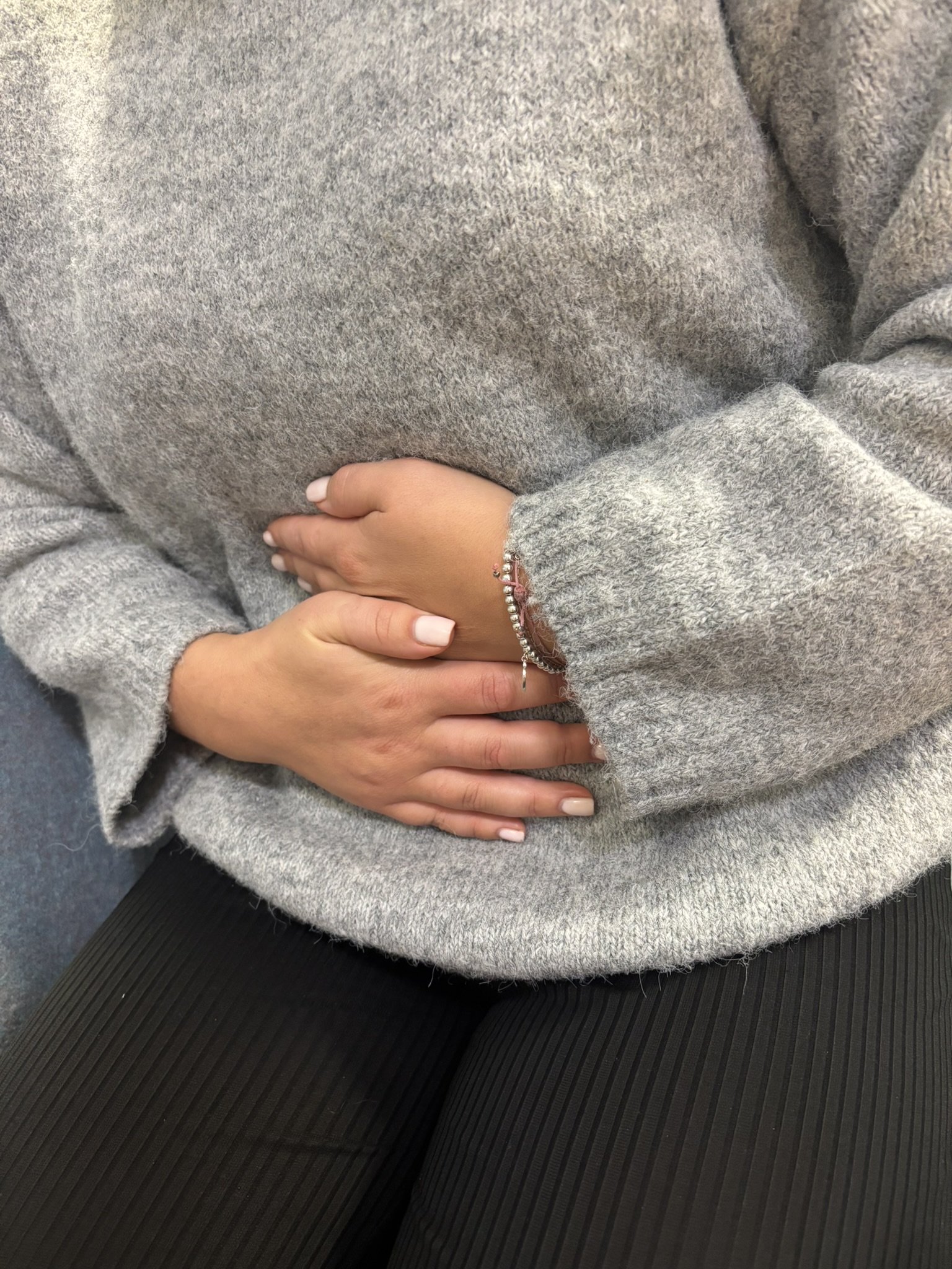 Person wearing a grey sweater and black pants, sitting on a black textured surface with hands crossed on their lap, wearing a pink bracelet with silver accents.