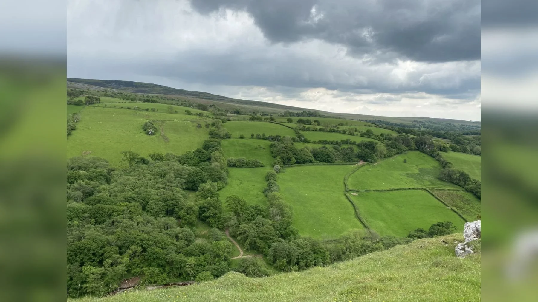 A green countryside landscape with rolling hills, fields separated by stone walls, and trees under a cloudy sky.