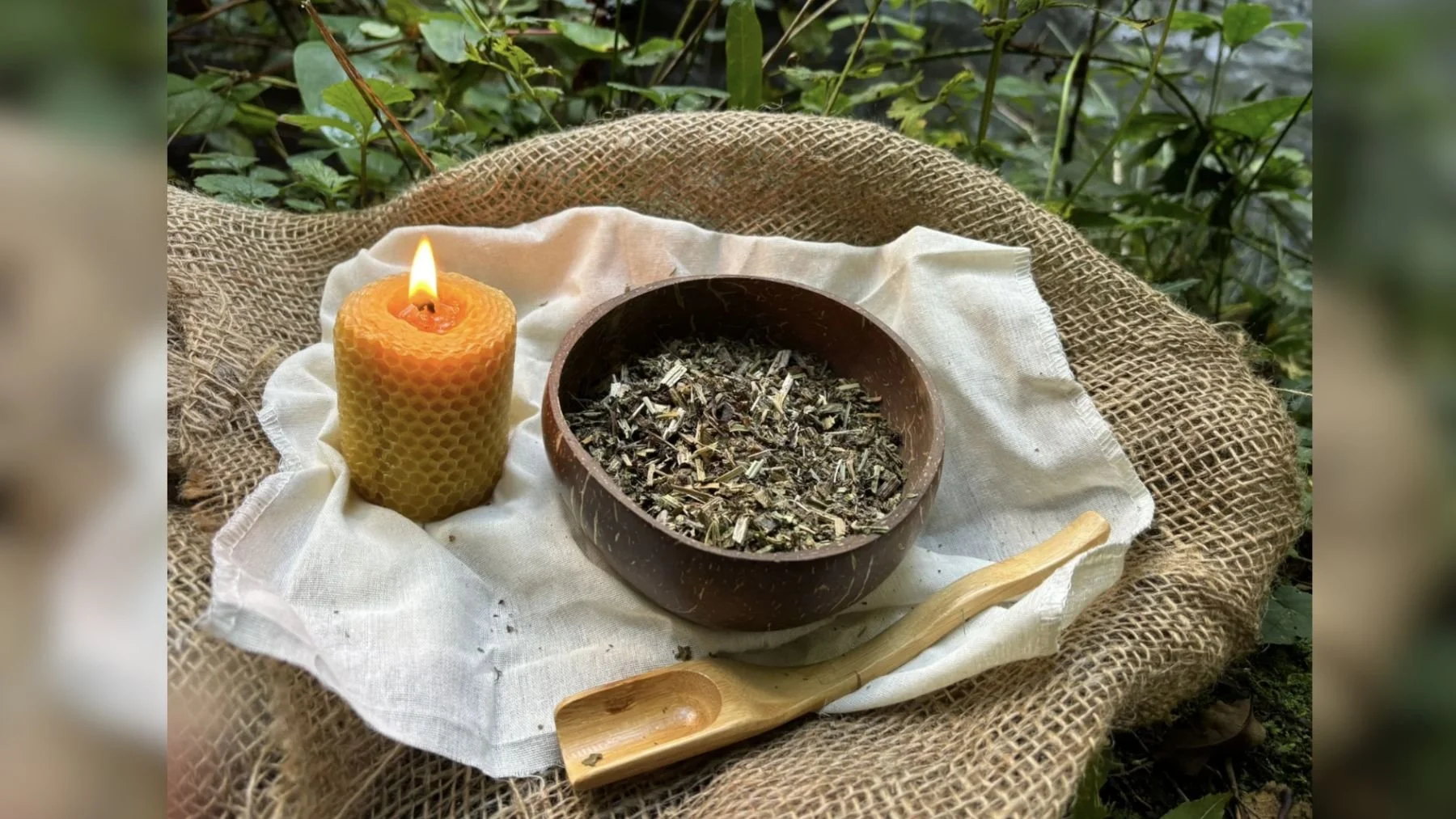 A lit beeswax candle, a wooden bowl filled with dried herbs and herbal mixture, and a wooden spoon on a white cloth, all on a burlap surface with green foliage in the background.