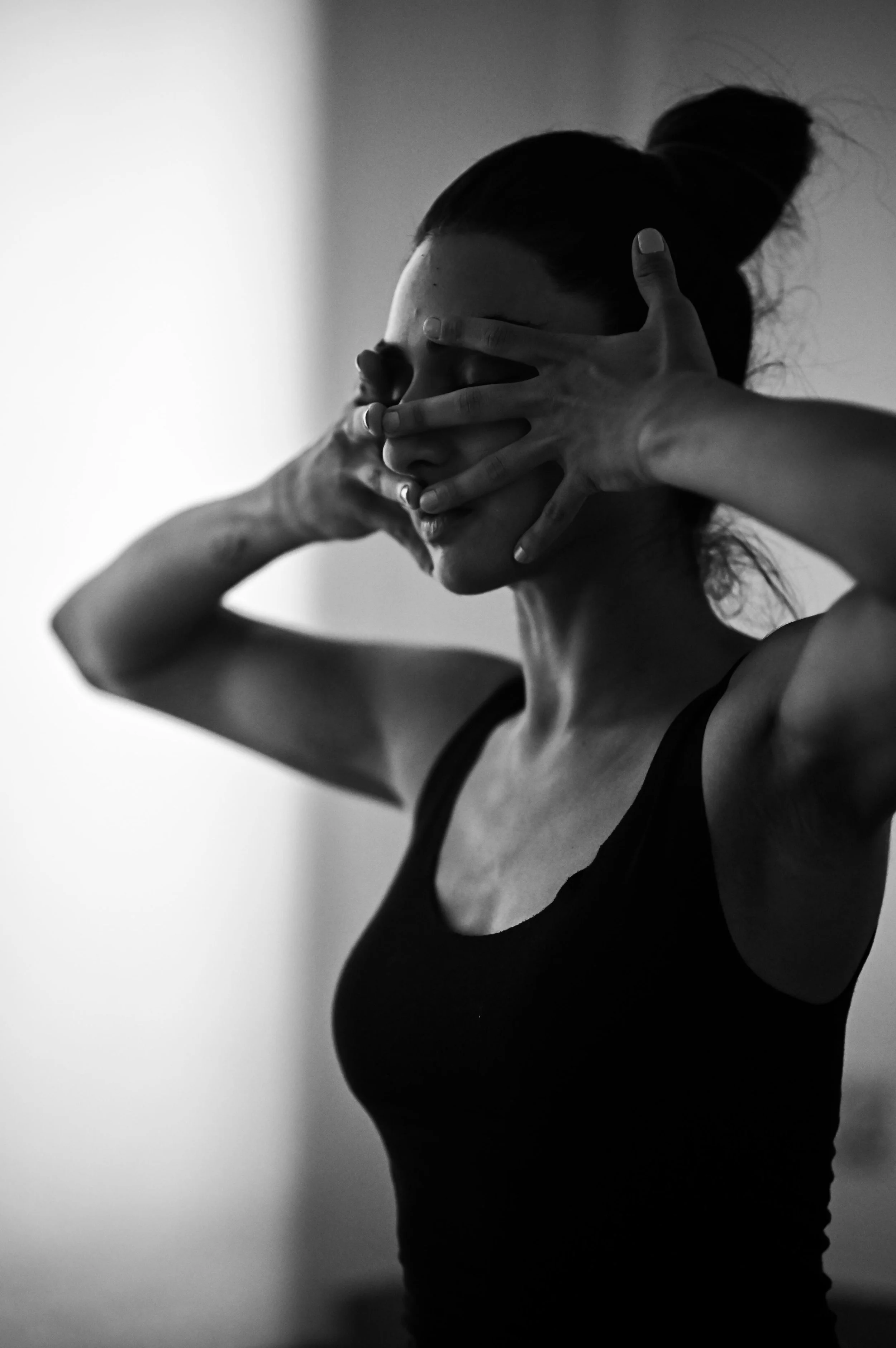 A woman with her hair in a bun and wearing a sleeveless top covering her face with her hands, fingers spread wide apart in a pose, black and white photograph.