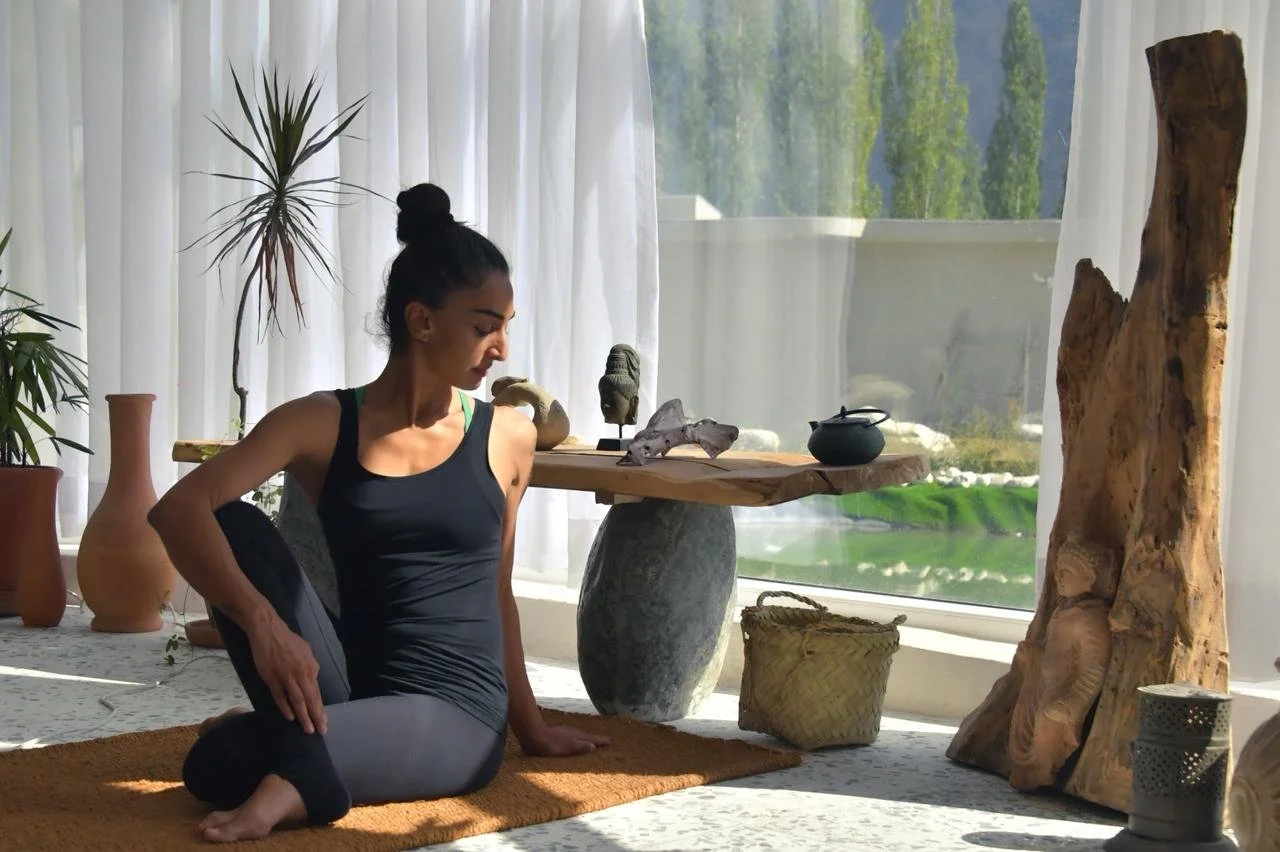 A woman practicing yoga in a cozy living room with neutral tones, surrounded by decorative vases and a stone fireplace, with soft lighting and white curtains.