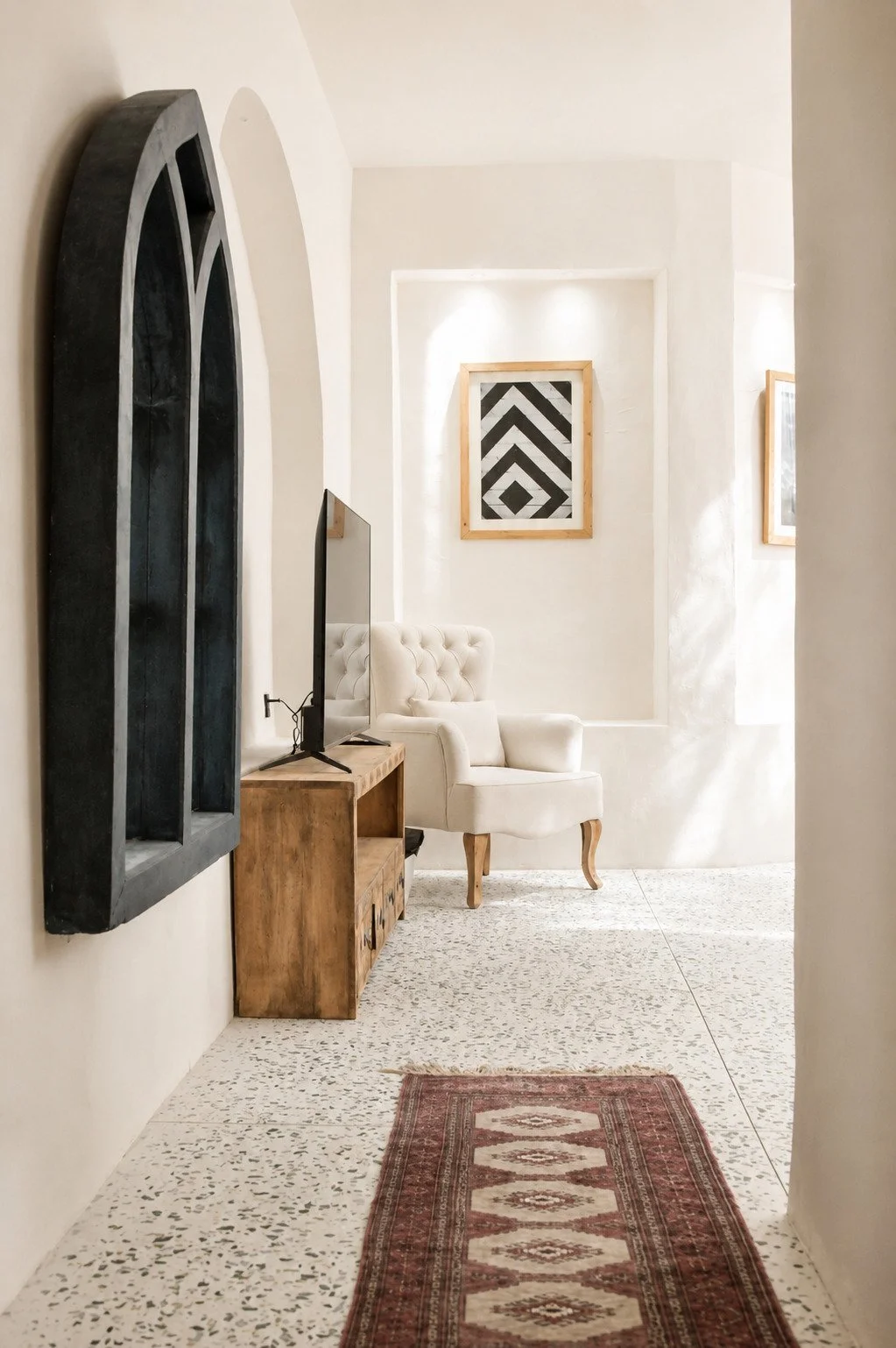 Living room with white armchair, wooden TV stand with TV, framed black-and-white geometric art, terrazzo flooring, and a red patterned rug.