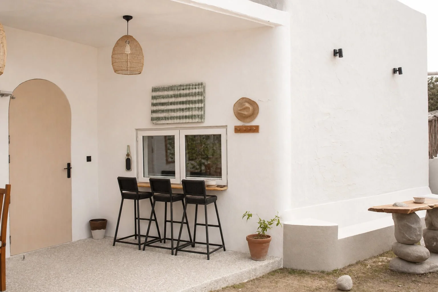 An outdoor patio area with a white stucco wall, three black bar stools at a wooden counter with a window behind, a woven hat and a small painting on the wall, a plant in a pot, and a small table with stone bases outside.