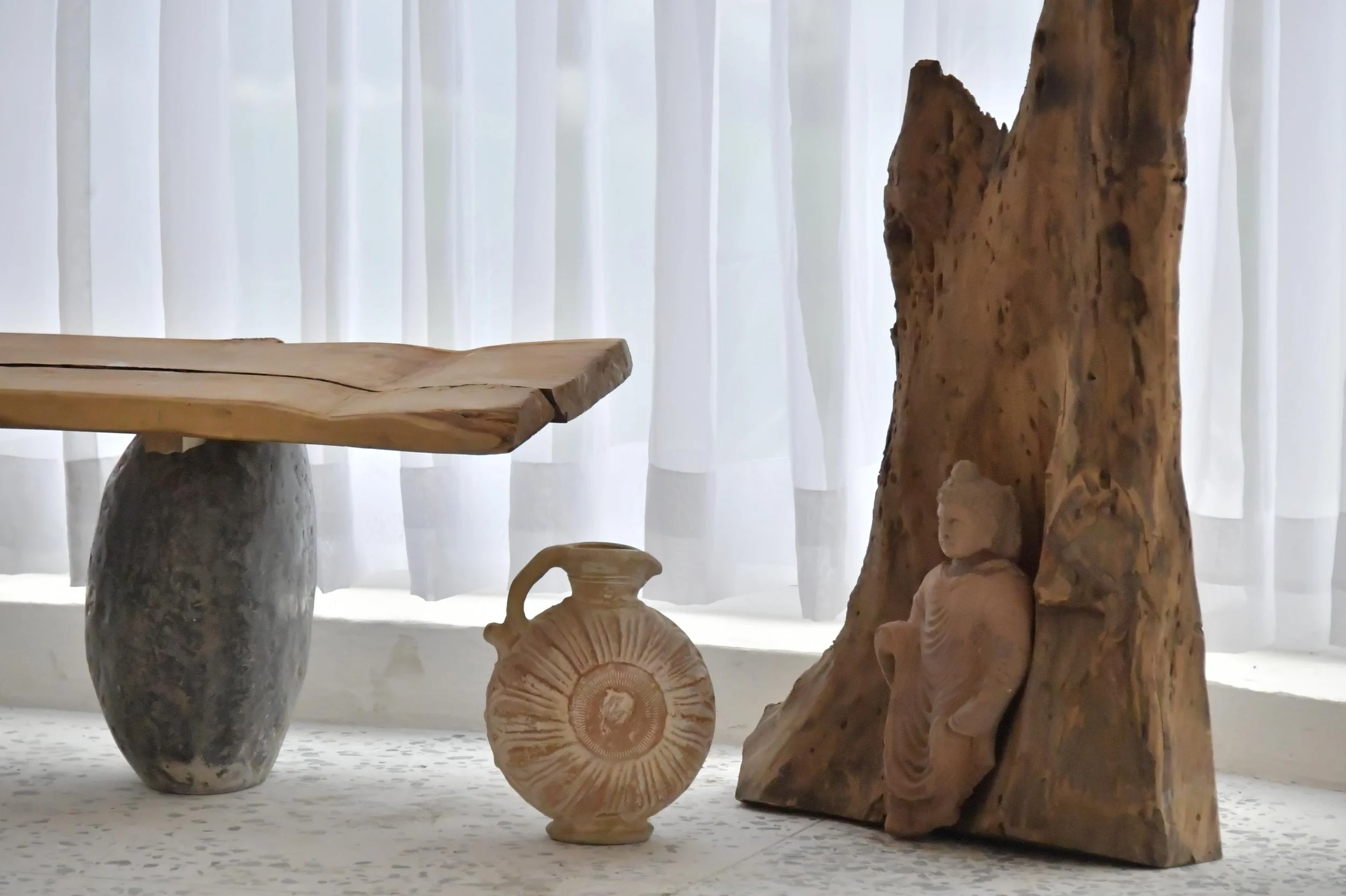 Various wooden and stone sculptures and objects on display in front of white curtains.