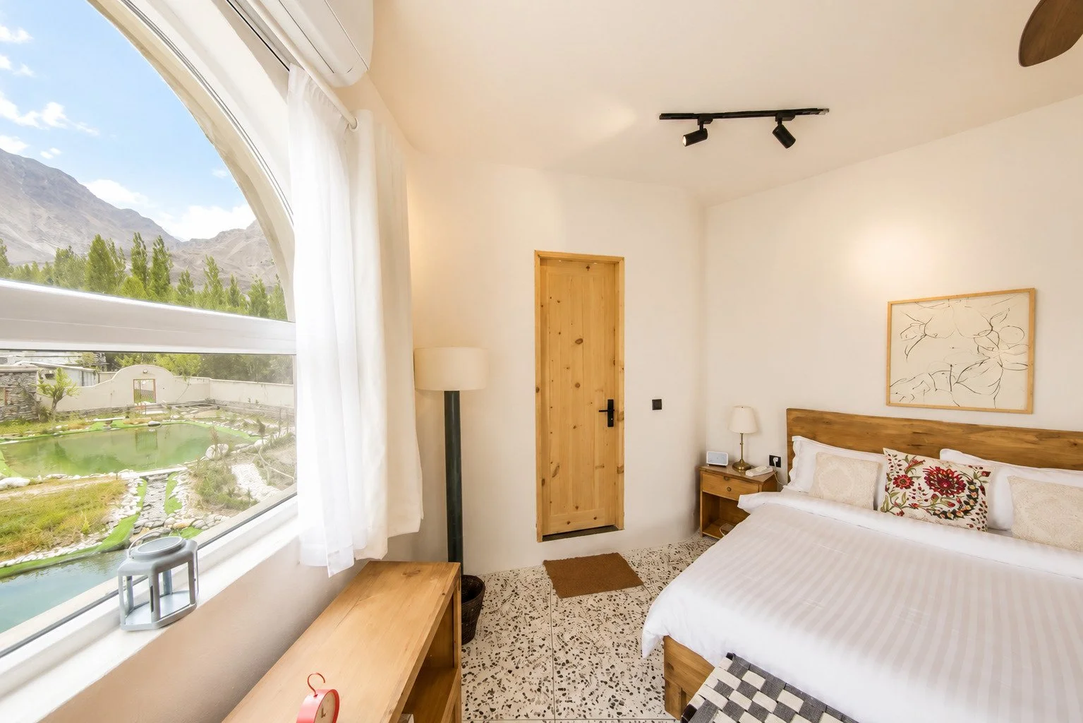 Bright hotel bedroom with white walls, wooden door, and bedside tables, one with a lamp and phone, a bed with floral and neutral pillows, large window with white curtains showing mountains and a garden outside, and a framed abstract artwork above the