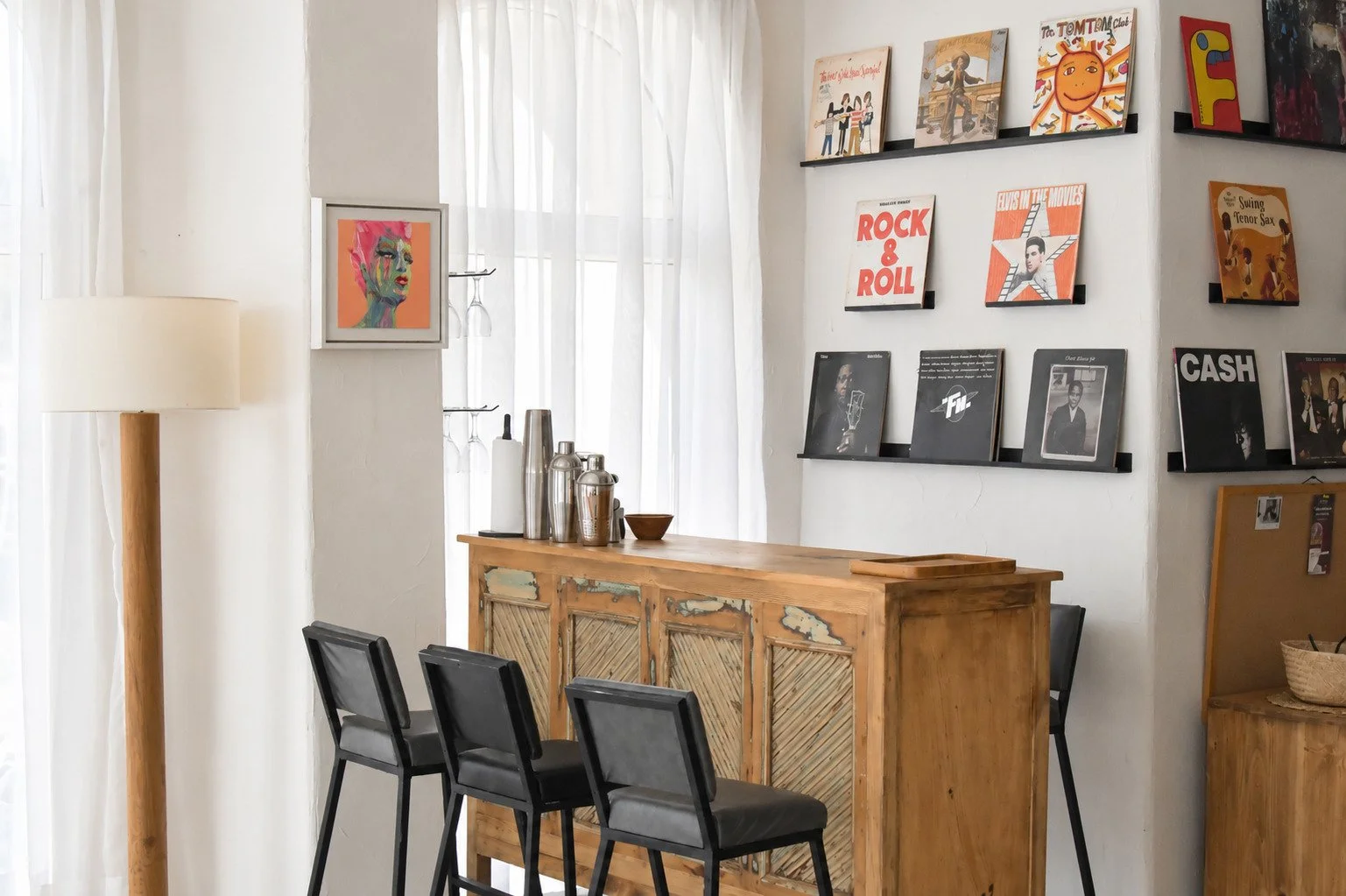 Interior view of a cafe or bar with a wooden counter, black chairs, a tall standing lamp, a shelf with vinyl records and album covers on the wall, and a window with sheer white curtains.