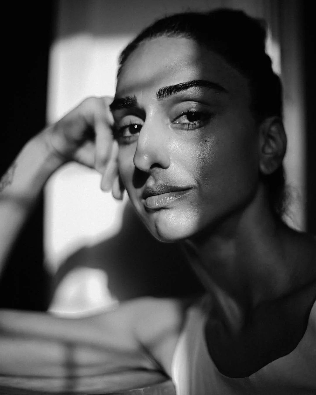 Close-up black and white portrait of a woman with dark hair, natural makeup, and a subtle smile, looking directly at the camera with her hand resting near her face.
