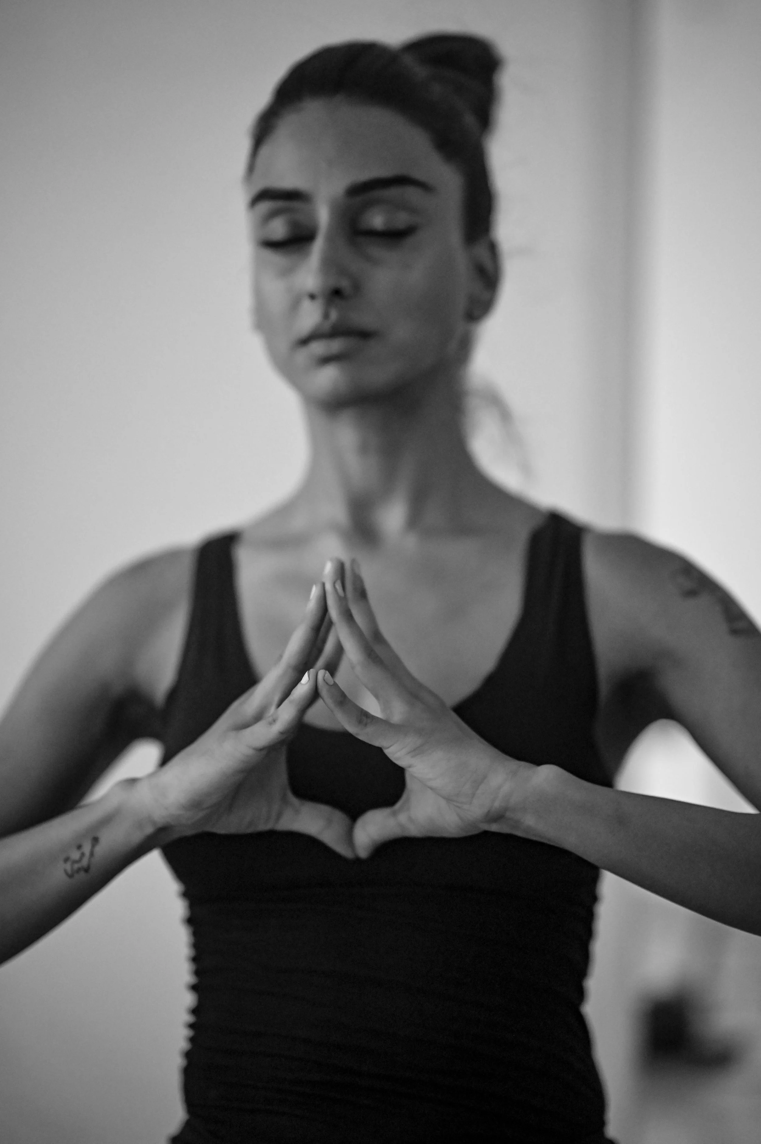 A woman with dark hair in a bun, wearing a black tank top, stands with her eyes closed, forming a heart shape with her hands.