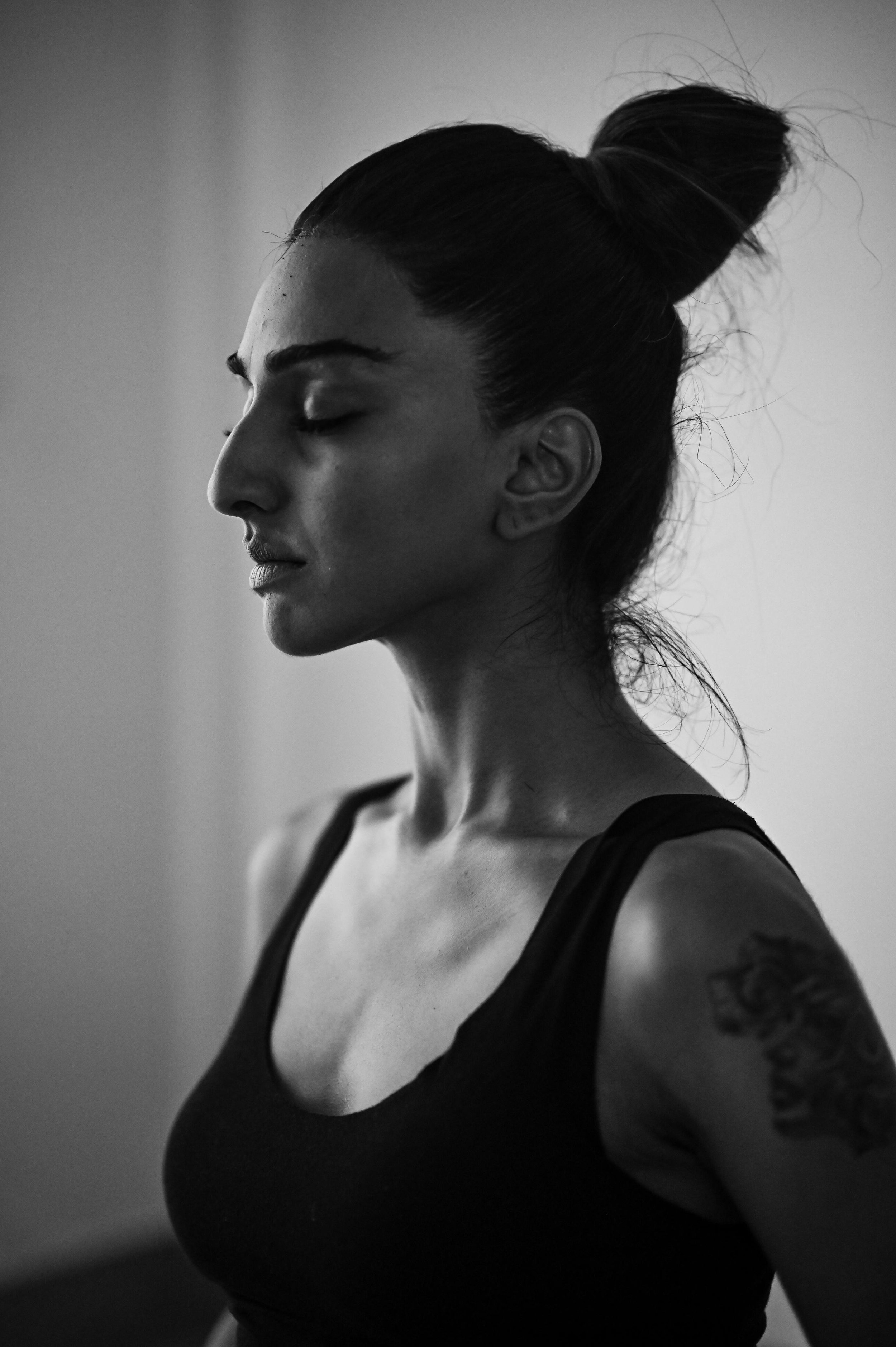 A black and white profile photo of a woman with her eyes closed, wearing a black tank top, with her hair styled in a high bun and a tattoo on her arm.
