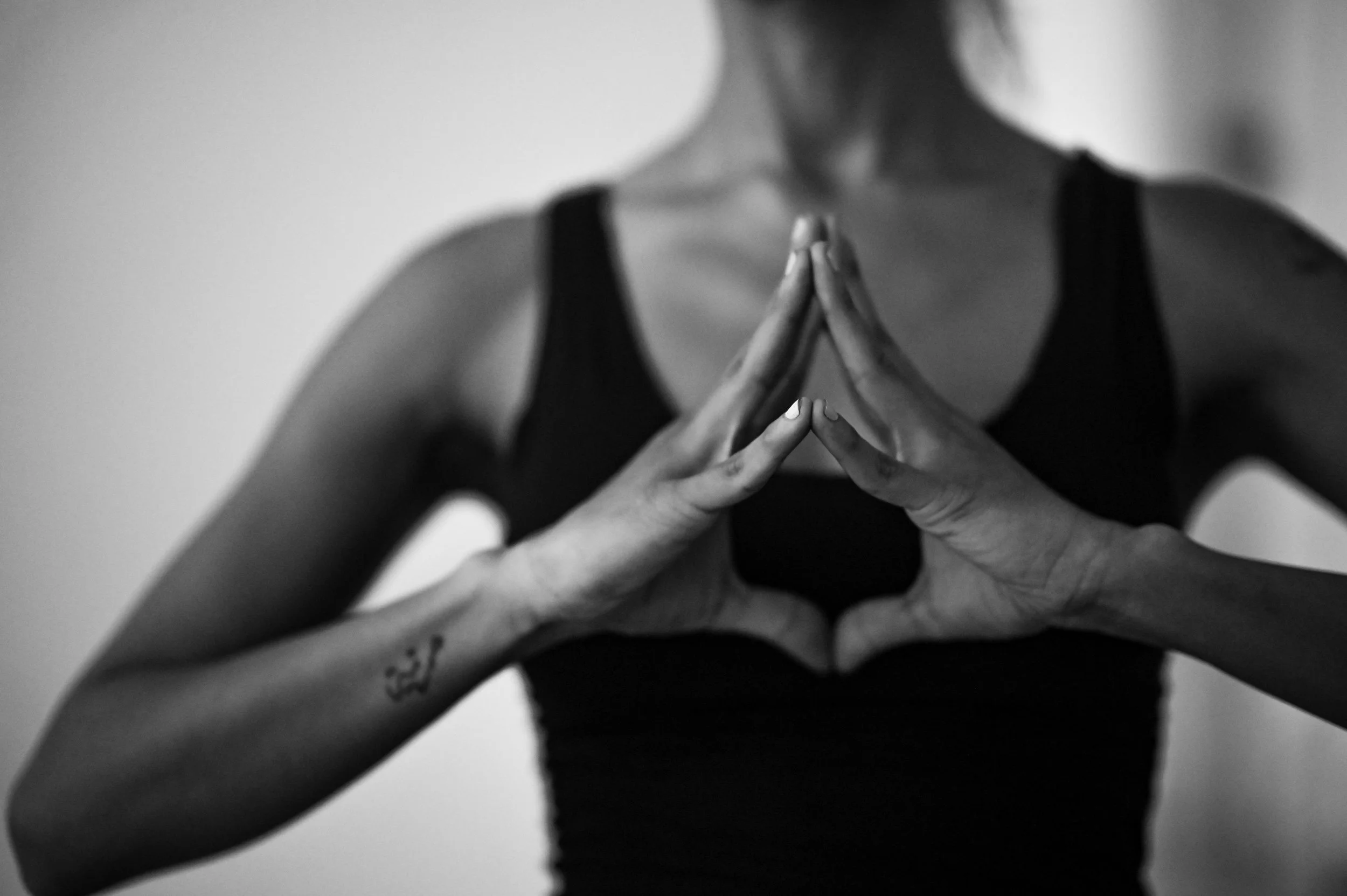 A woman practicing yoga or meditation, standing in a black tank top, with her hands in a prayer position in front of her chest.