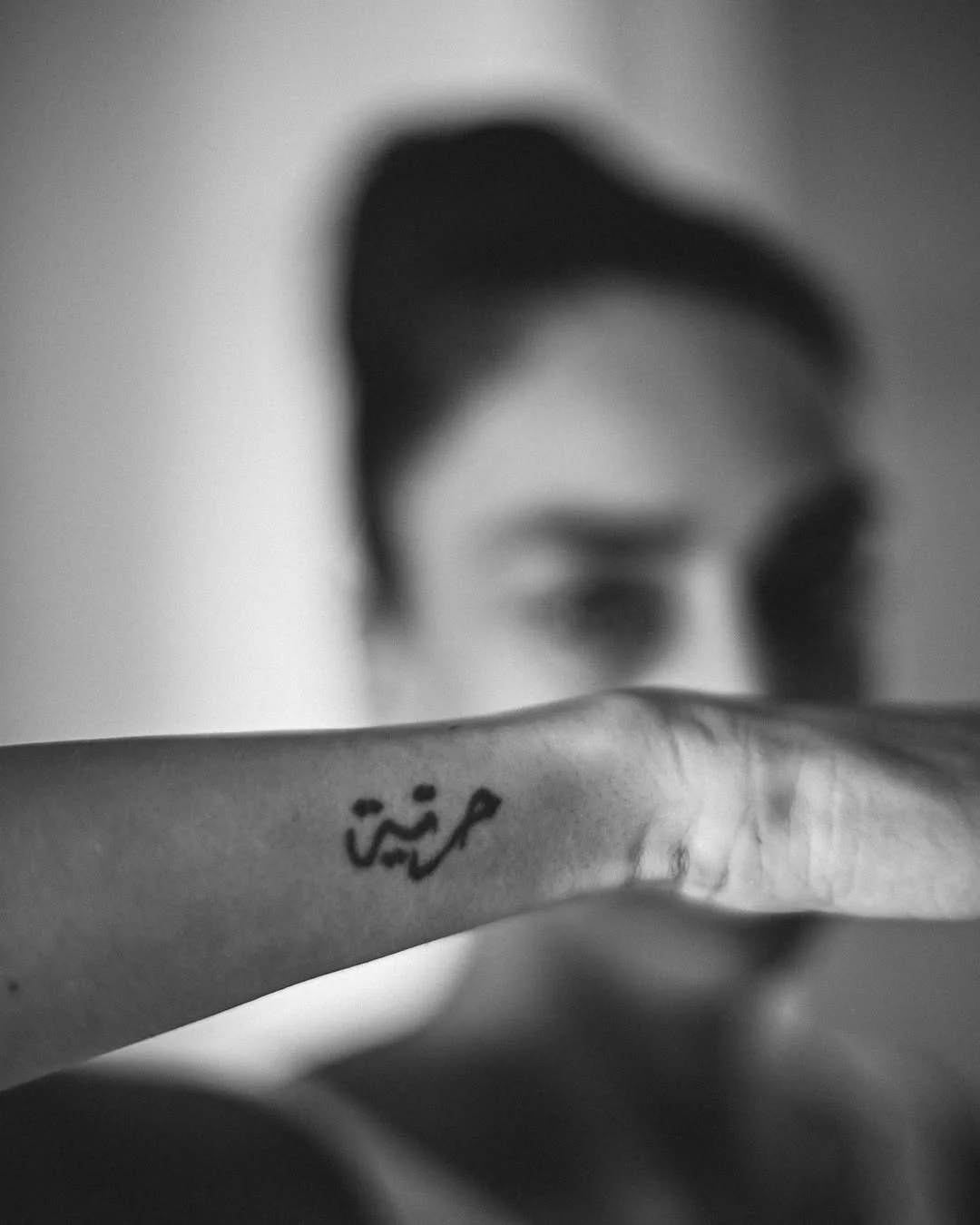 A person with dark hair, partially visible face, holding their arm with Arabic script tattooed on it, in black and white photo.