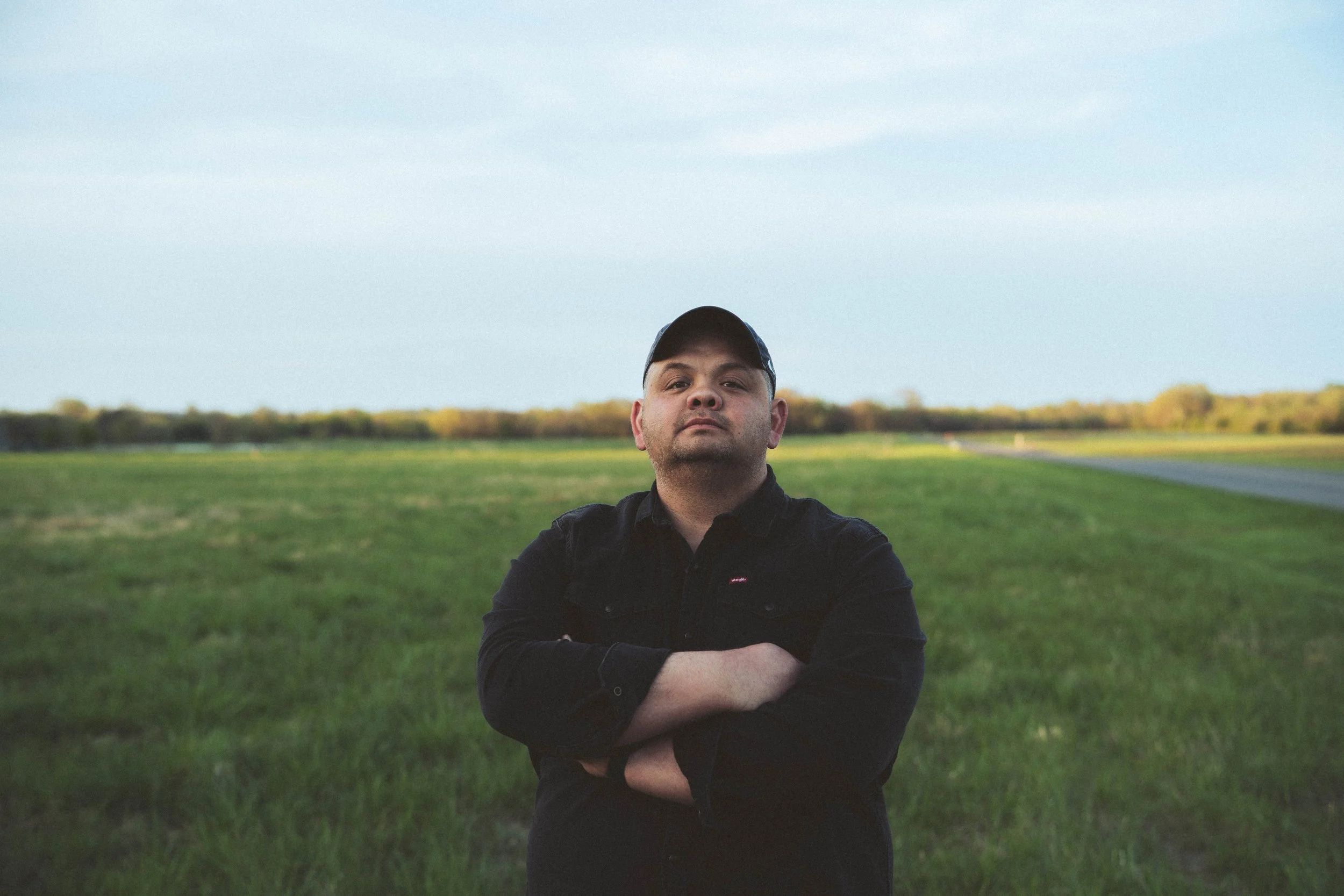 Phillip Bowen stands in a field with his arms crossed wearing a black button up shirt and black ball cap.
