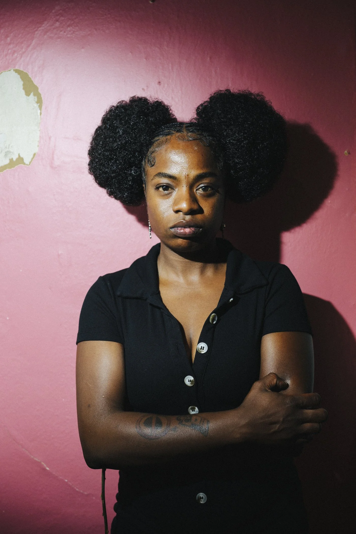 Sunny War stands with a serious look on her face, with arms crossed against a dark pink wall.