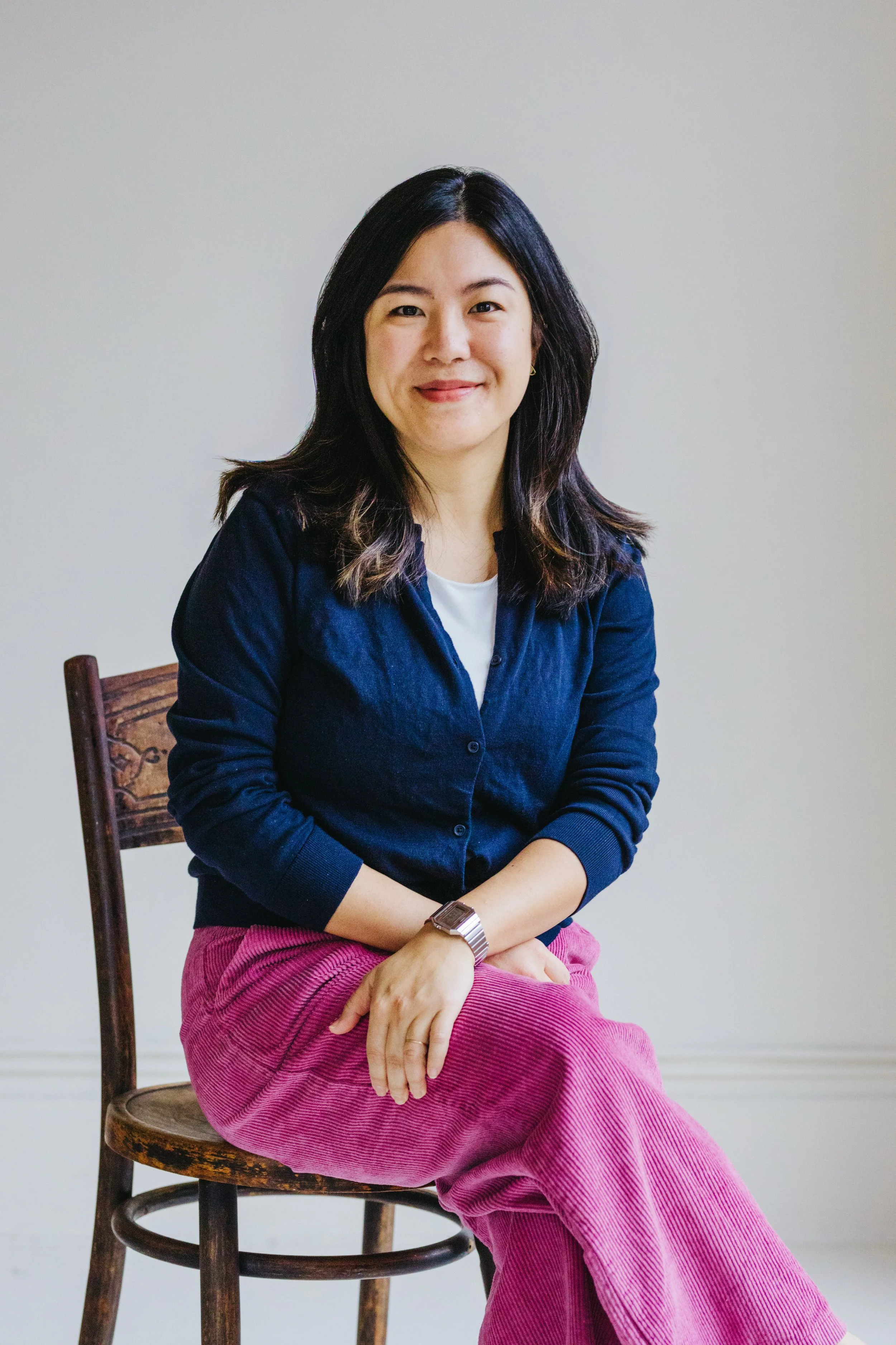 A woman with shoulder-length dark hair sitting on a wooden chair against a plain light gray wall, smiling, wearing a dark blue cardigan, a white undershirt, a wristwatch, and pink corduroy pants.
