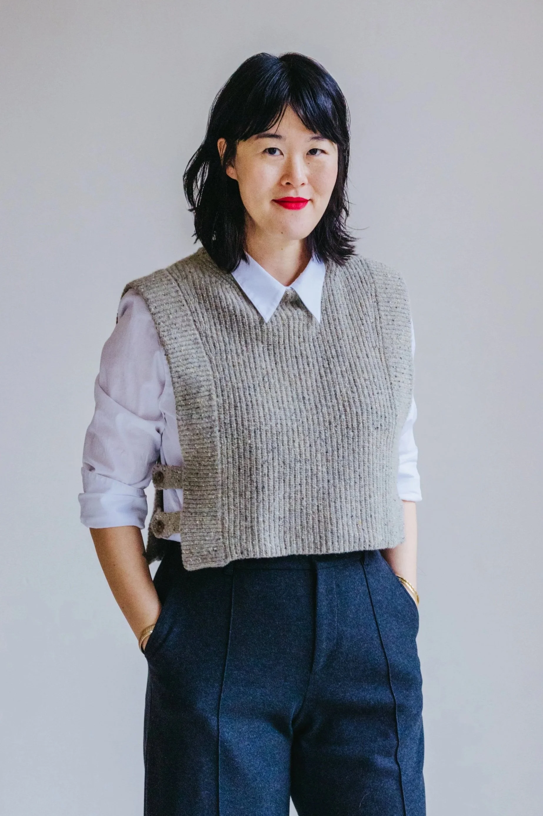 A woman with black hair, wearing a white shirt with rolled-up sleeves, a gray knitted vest, and high-waisted charcoal pants, standing against a plain light gray background.