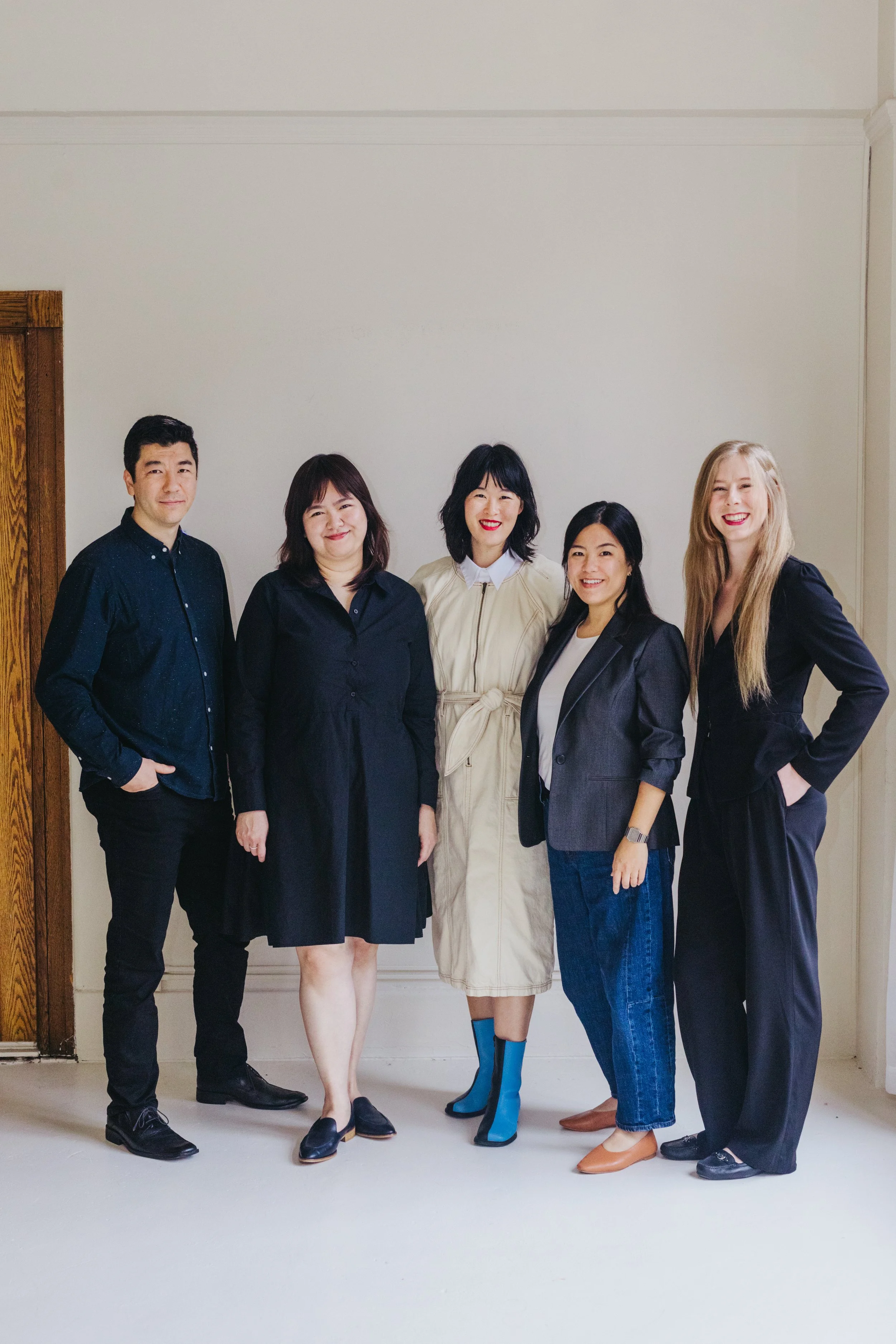 Group of six diverse individuals standing together inside a room, smiling for the camera. They are dressed in casual and semi-formal attire, with some wearing jackets and others in dresses or skirts.