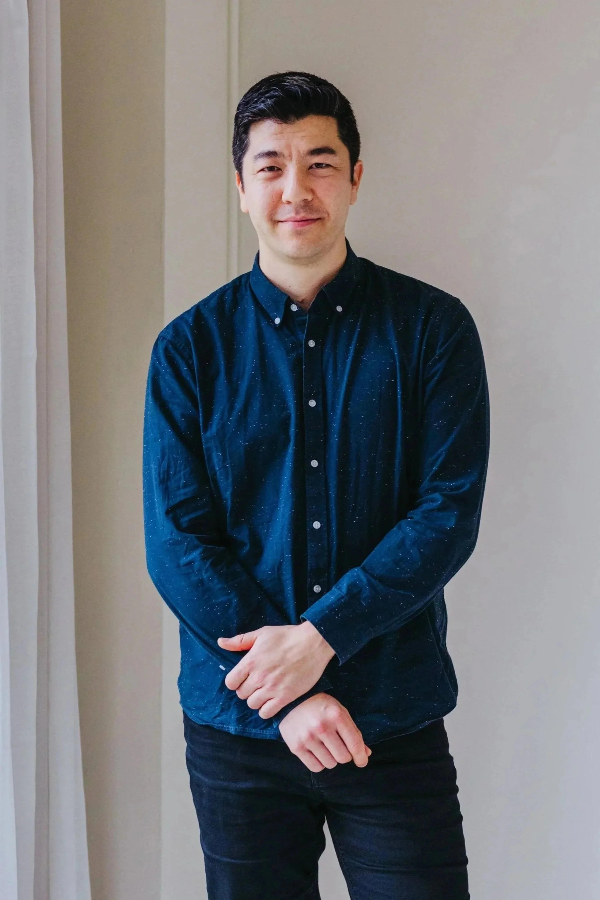 A man with short dark hair, wearing a dark blue button-up shirt, standing against a neutral-colored wall. He has a slight smile and is crossing his hands in front of him.