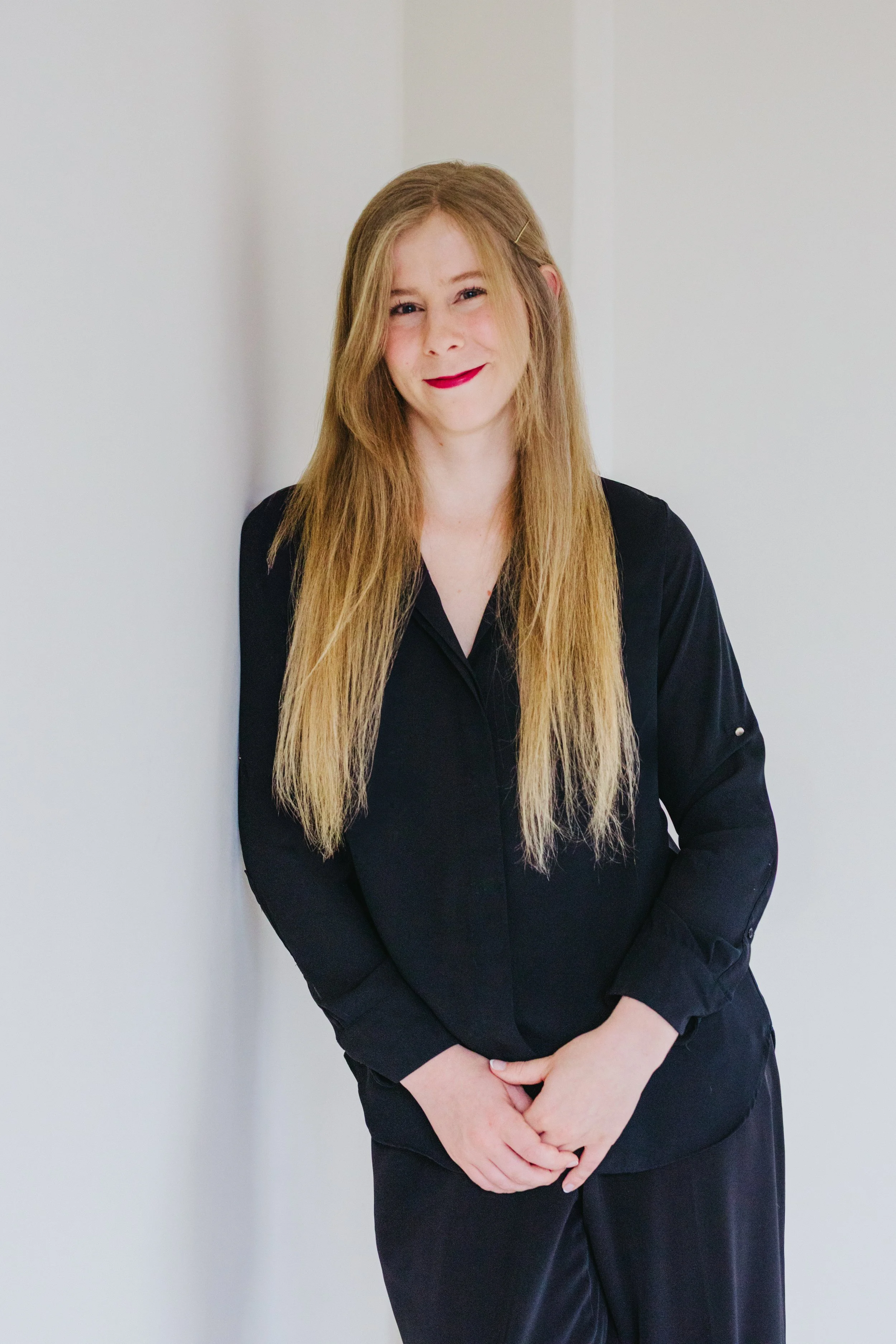 A woman with long blonde hair, wearing a black shirt, standing against a white wall, smiling gently, with her hands clasped in front of her.