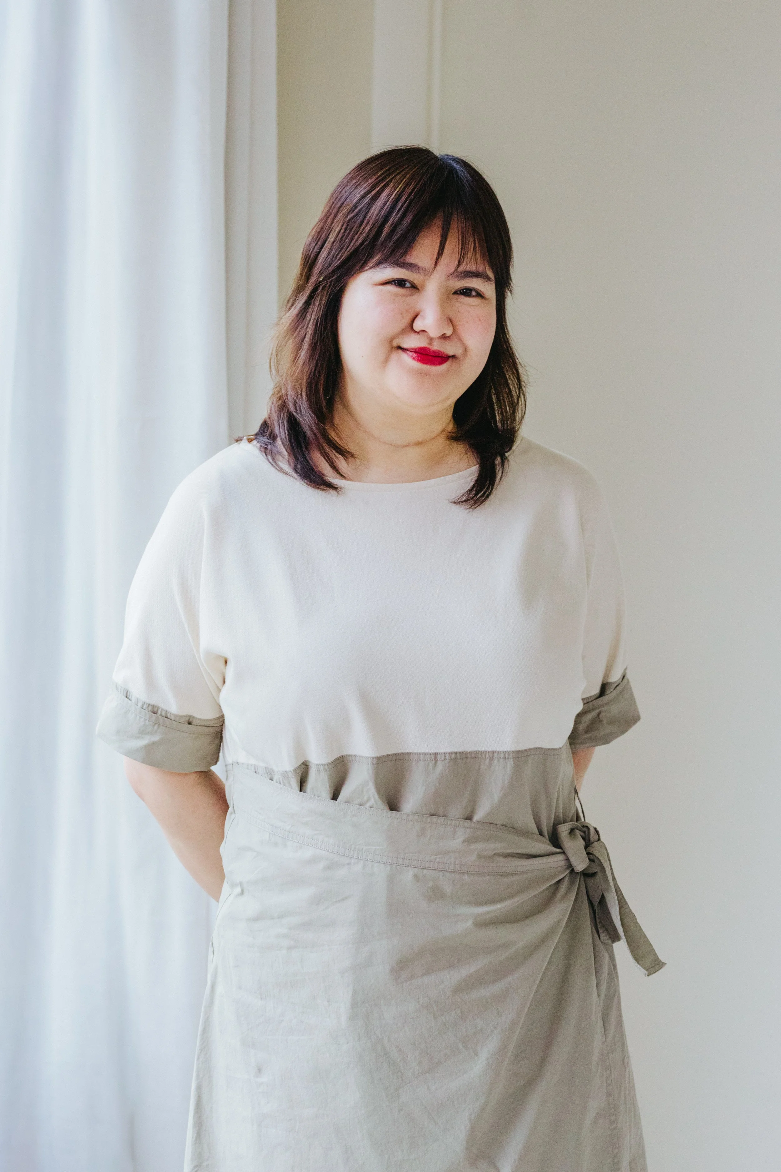 A woman with shoulder-length dark hair and red lipstick standing indoors near a light-colored wall and sheer curtains, wearing a beige and white dress with a tied waist.