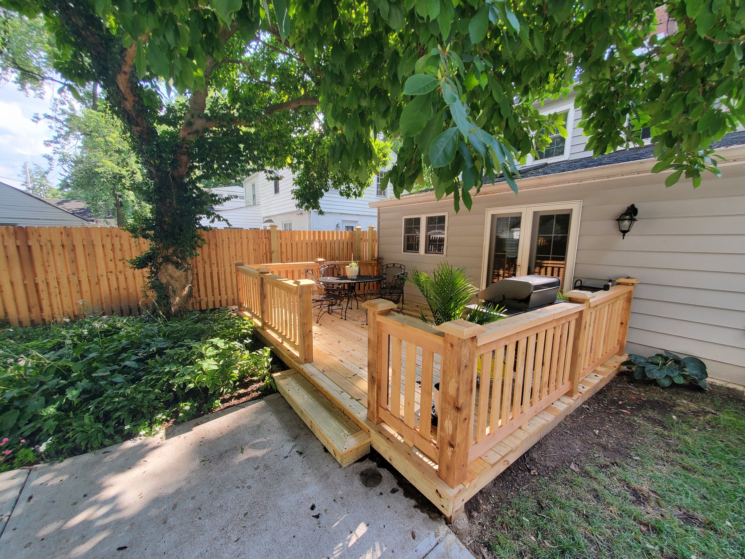 A newly built wooden deck attached to a house, with a railing, outdoor furniture, a grill, and surrounded by a garden with trees and plants.