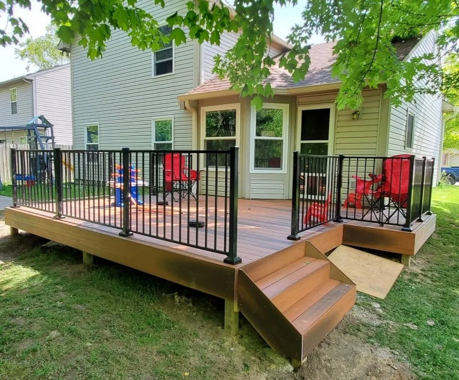 A raised wooden deck with black railing attached to a two-story house with white siding. The deck has two steps leading down to the yard and features red chairs, a small table, and a children's play set with a slide.