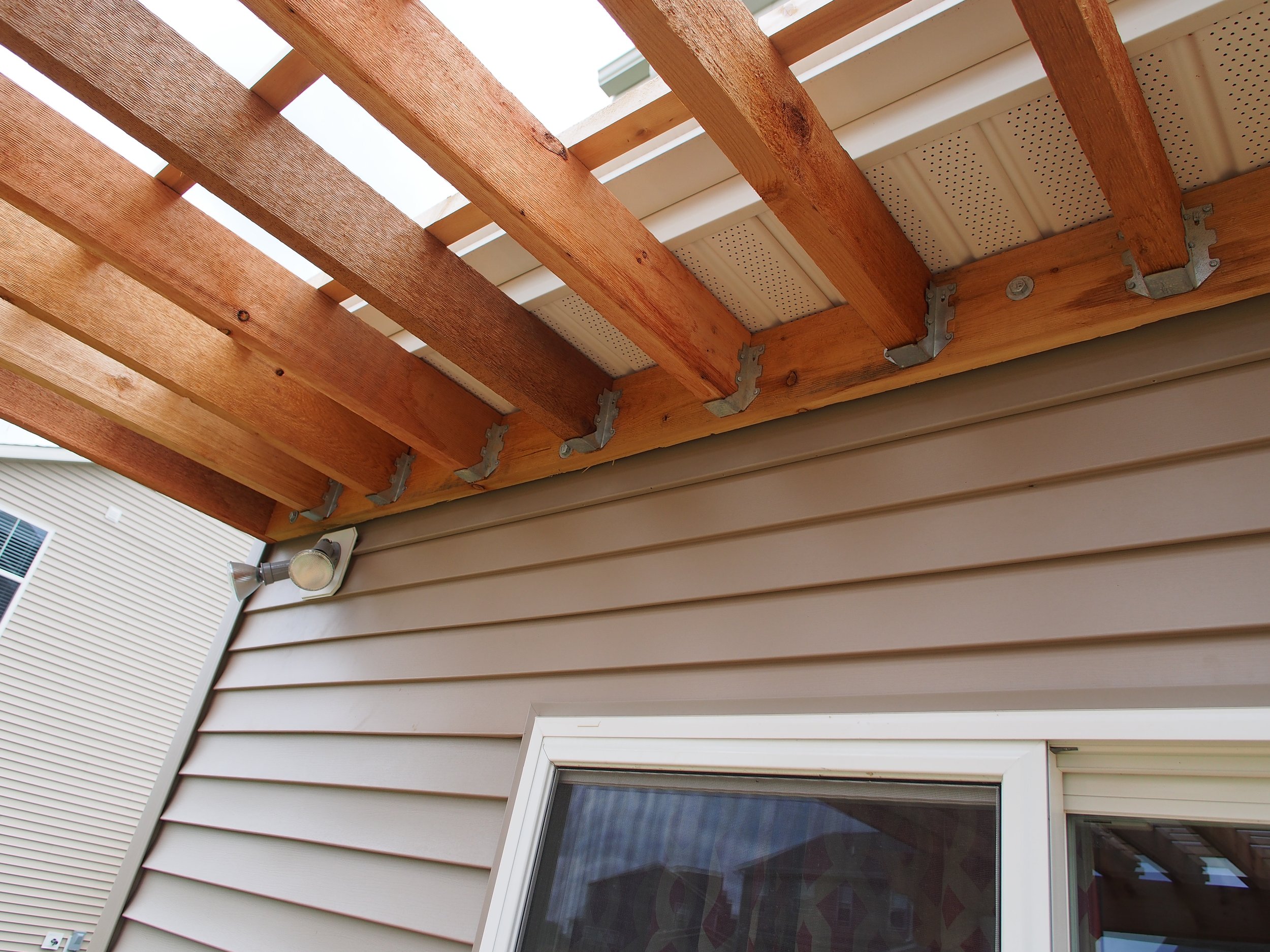 View of a wooden deck structure attached to the exterior of a house, with a beige vinyl siding wall and a window below, a security light on the wall, and part of a neighboring house visible in the background.