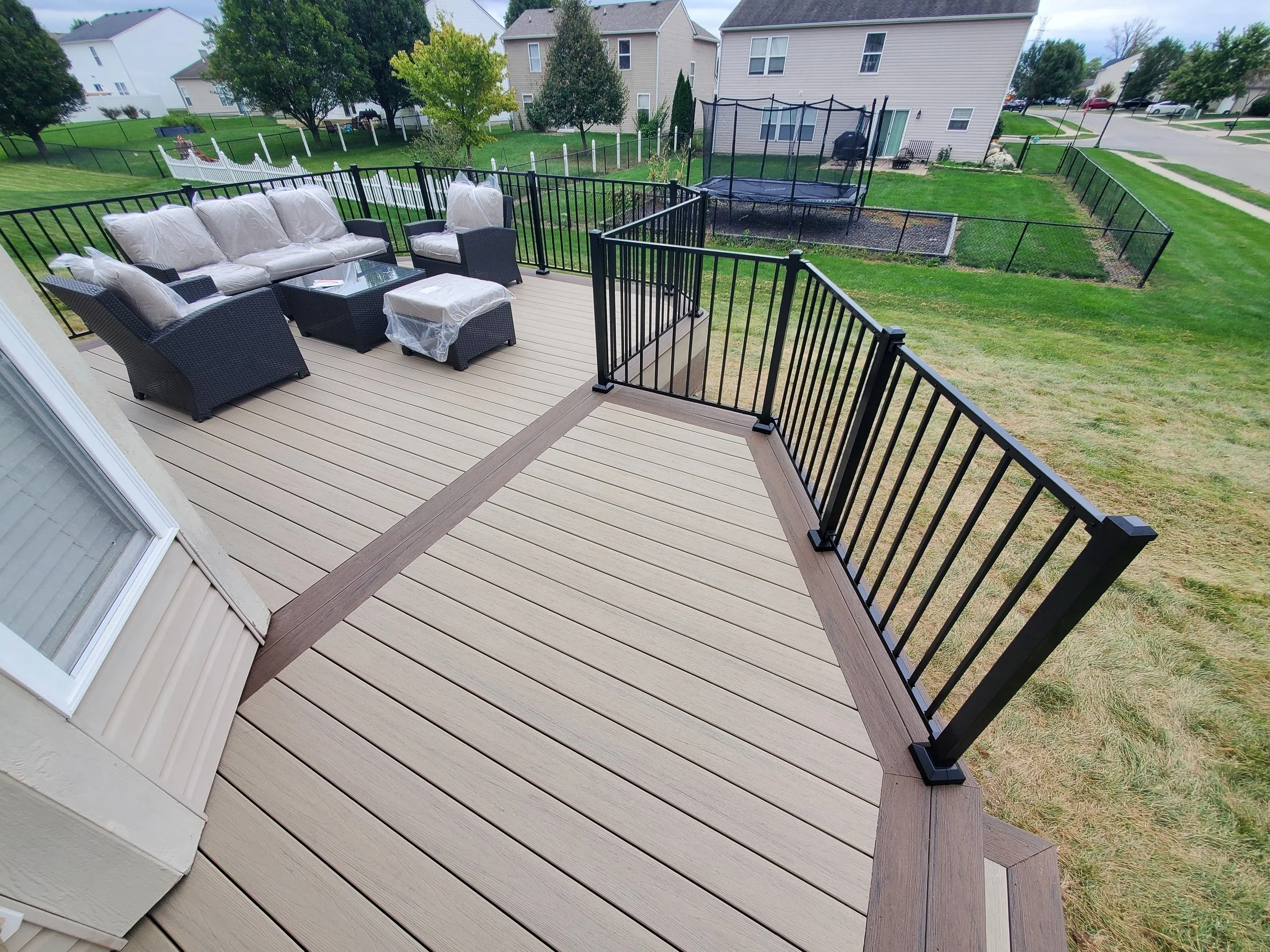 A new wooden deck with black metal railings, furnished with outdoor seating including a sofa, chairs, and a small table, overlooking a backyard with a trampoline and neighboring houses.