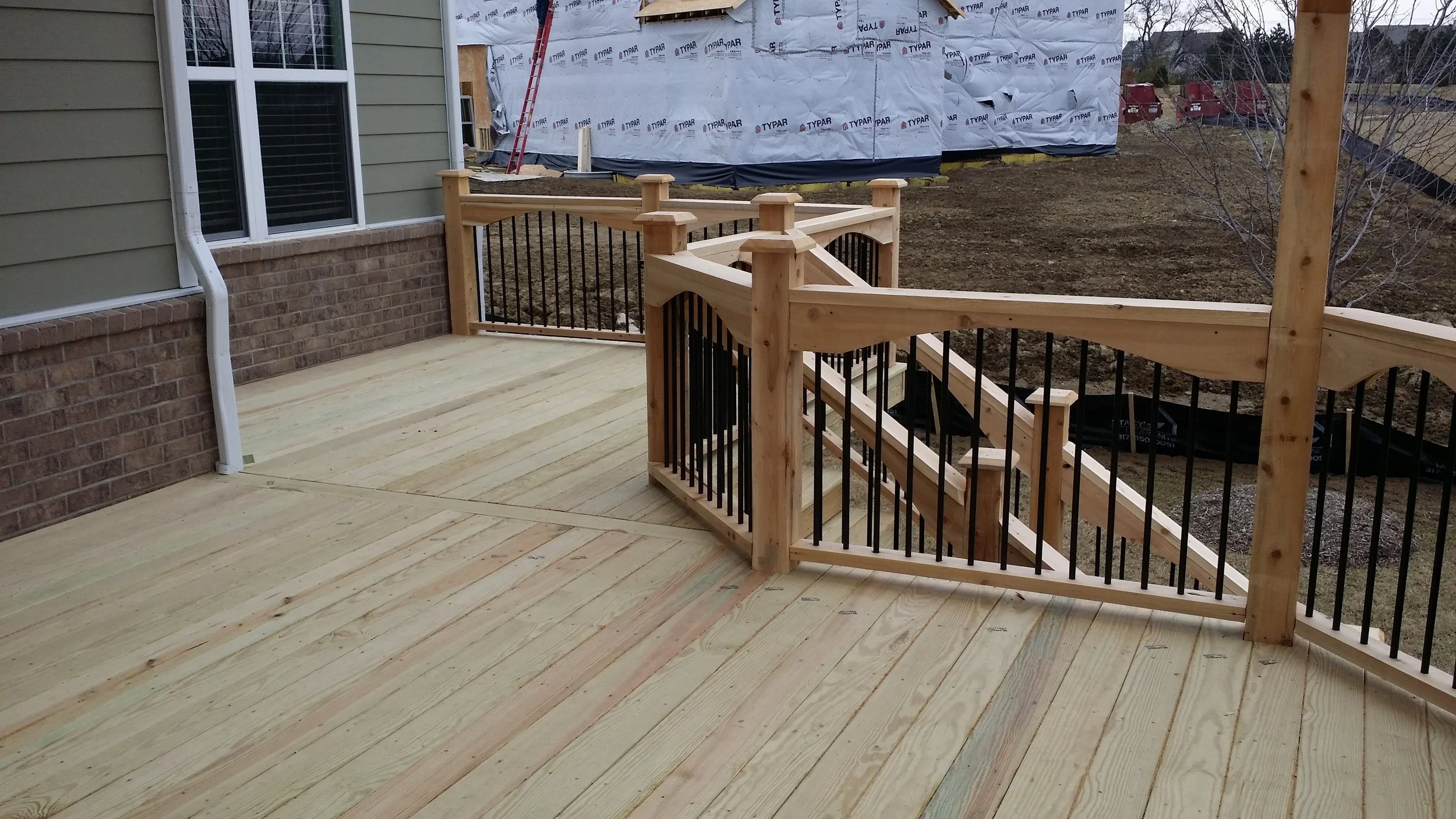 Photo of a newly built wooden deck with black metal railing, attached to a house with green siding and brick foundation. Stairs lead down to a yard under construction with scaffolding and a house in the background.