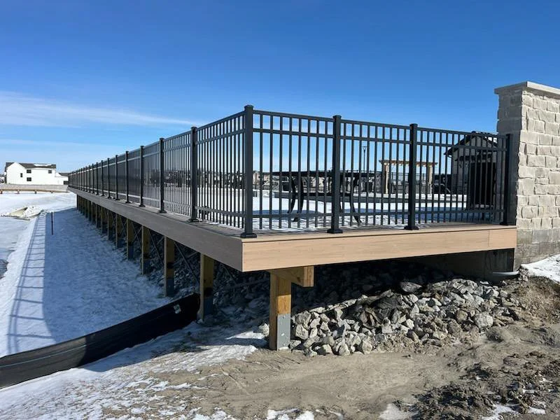Wooden deck with black metal railing on snow-covered ground in a suburban area, under a clear blue sky.