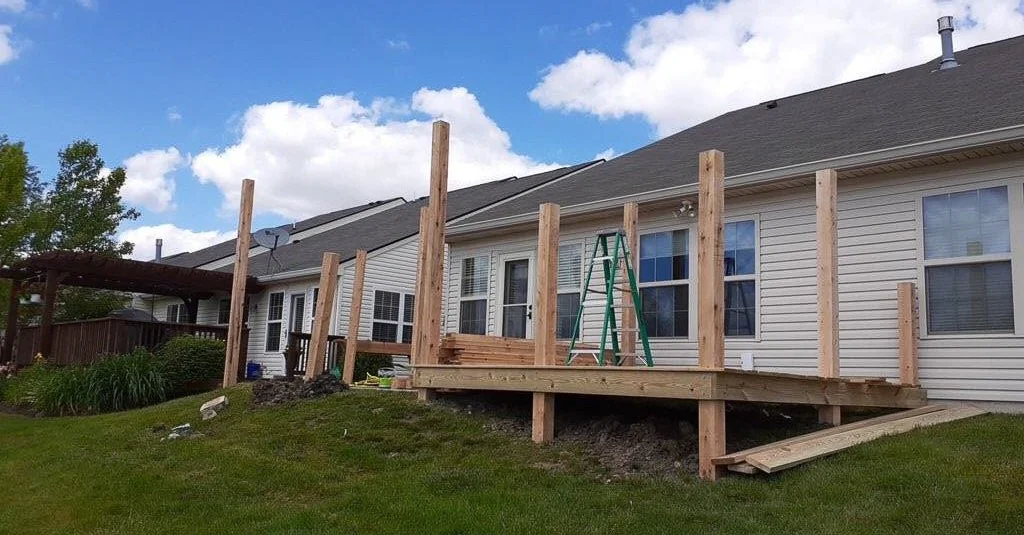 Construction of a wooden deck on the backyard of a house with installed posts and a green ladder, with a blue sky and some clouds overhead.