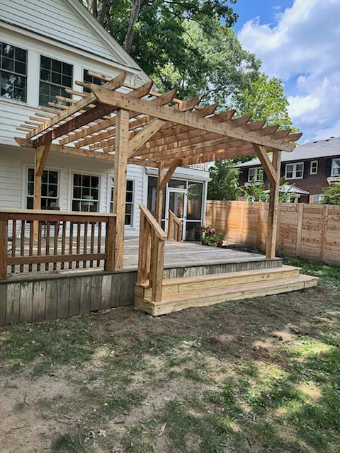 New wooden deck with pergola in backyard of a house, with stairs leading up to it and a railing on one side.