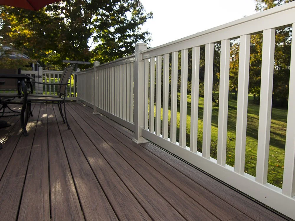 Image of a wooden deck with a white railing, an outdoor dining table, and chairs, with trees and a grassy yard in the background.