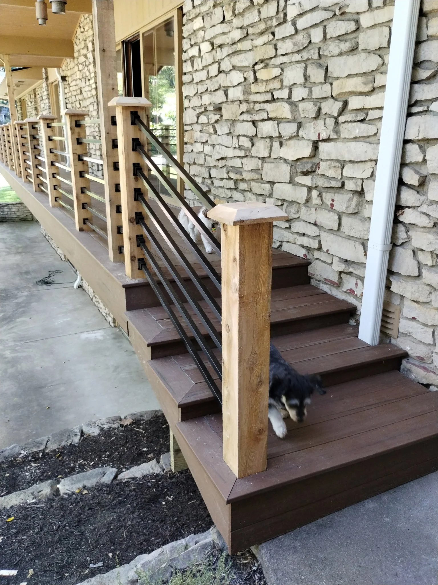 A house outdoor porch with a wooden deck, black metal railing, stone wall, and a puppy walking down the steps.
