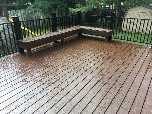 Wet wooden deck with a built-in wooden bench, surrounded by a black metal railing and a backyard with trees and a shed.