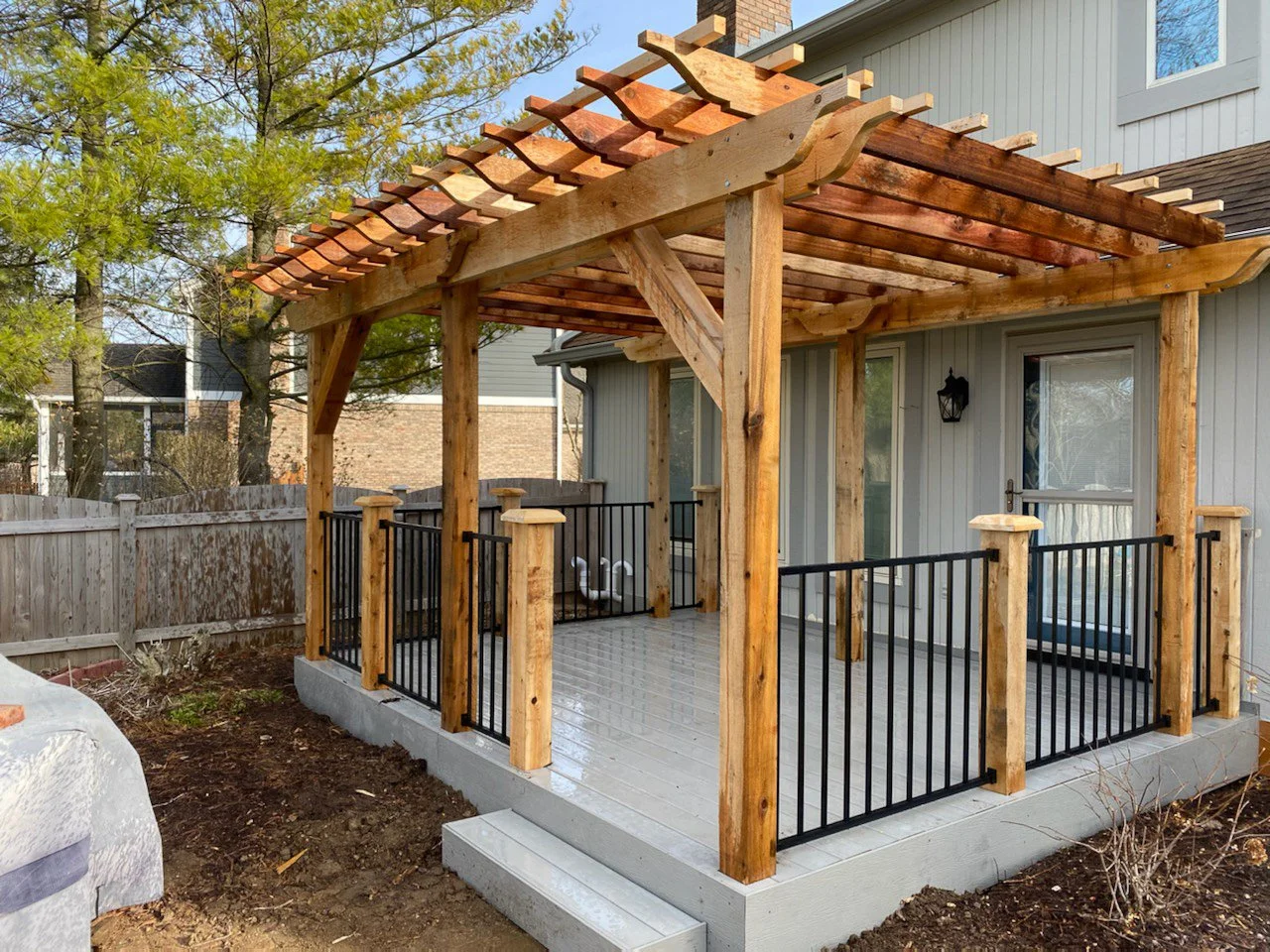 Backyard patio with a wooden pergola with decorative curved rafters, black metal railing, and light gray painted wooden floor, attached to a house with sliding glass door and exterior wall light.