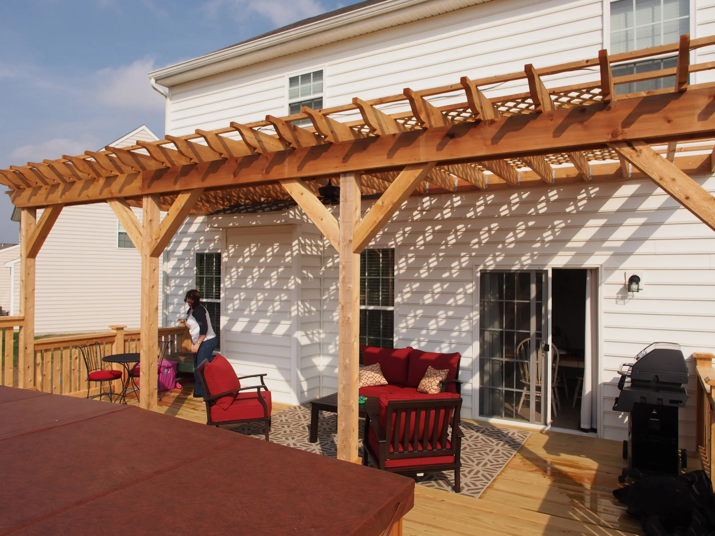 Backyard deck with a wooden pergola, red outdoor furniture, and a person standing near a small table with chairs.