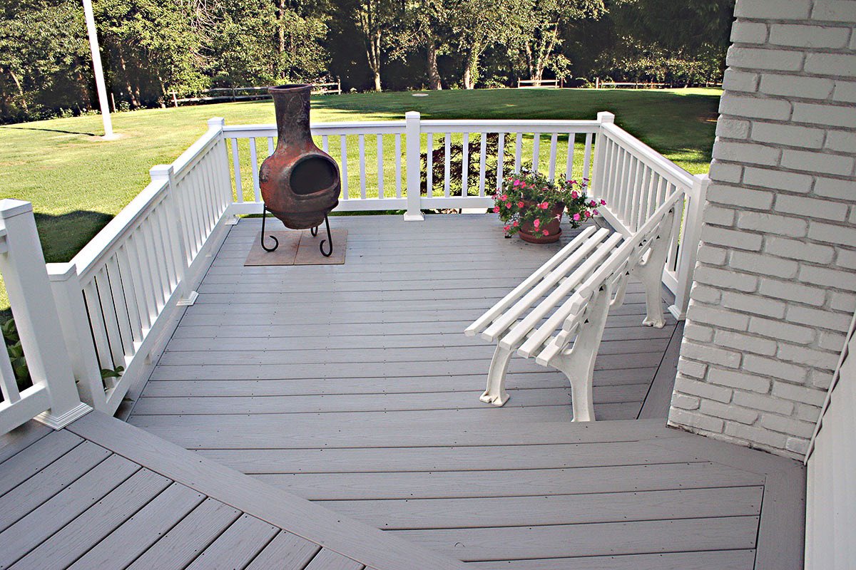 A small outdoor deck with white railing, a white bench, a large clay chiminea, and a potted plant with pink flowers, overlooking a green lawn and trees.