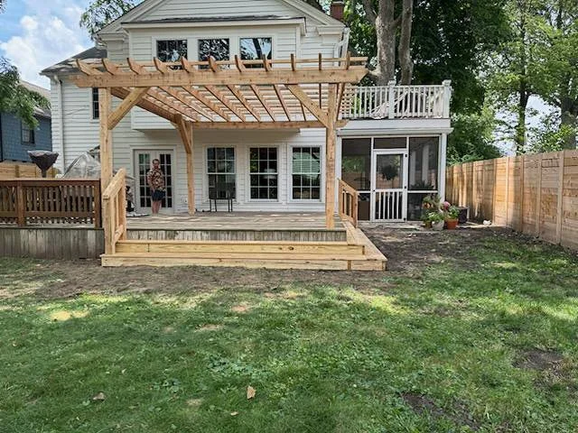A backyard with a house featuring a wooden deck and pergola in progress. The deck has a small set of stairs, and there are plants in pots near the house. A person is standing near the house on the deck, and the yard has green grass with some patches 