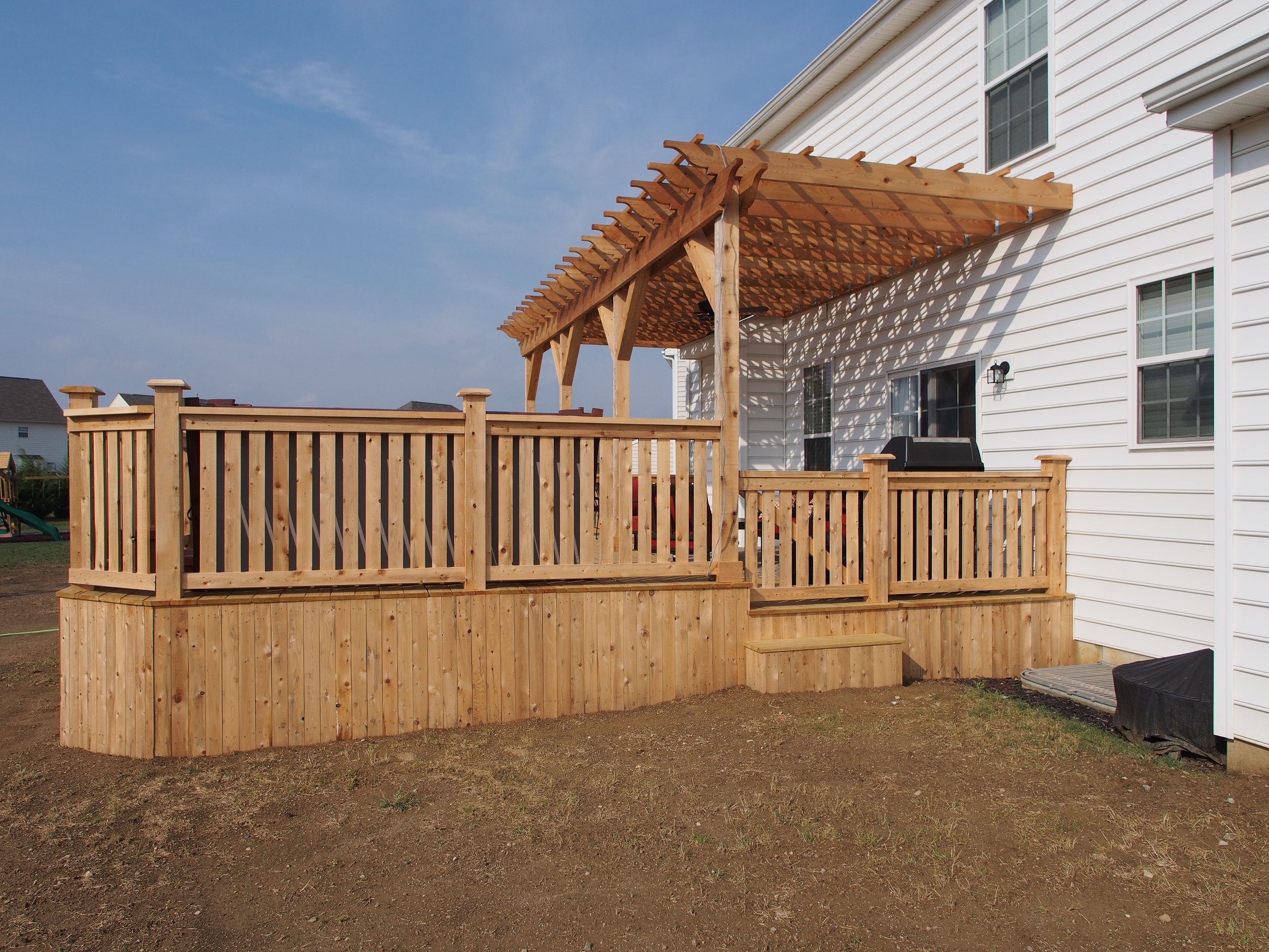 Newly built wooden deck with railing and pergola attached to a white house, with outdoor space and neighboring houses in the background.
