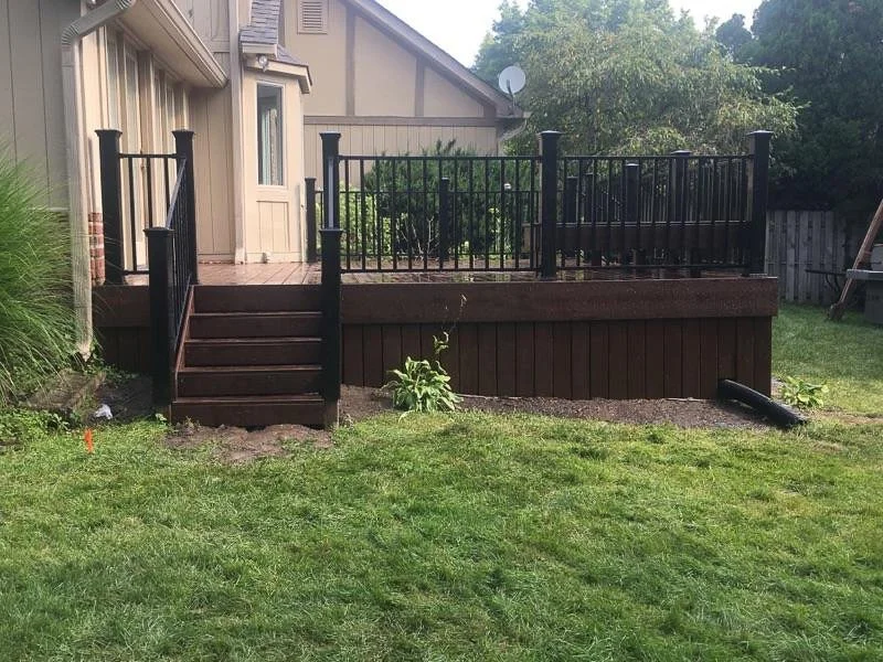 A raised wooden deck with black metal railing in a backyard, accessed by stairs from the green lawn, with surrounding trees and a neighboring house.