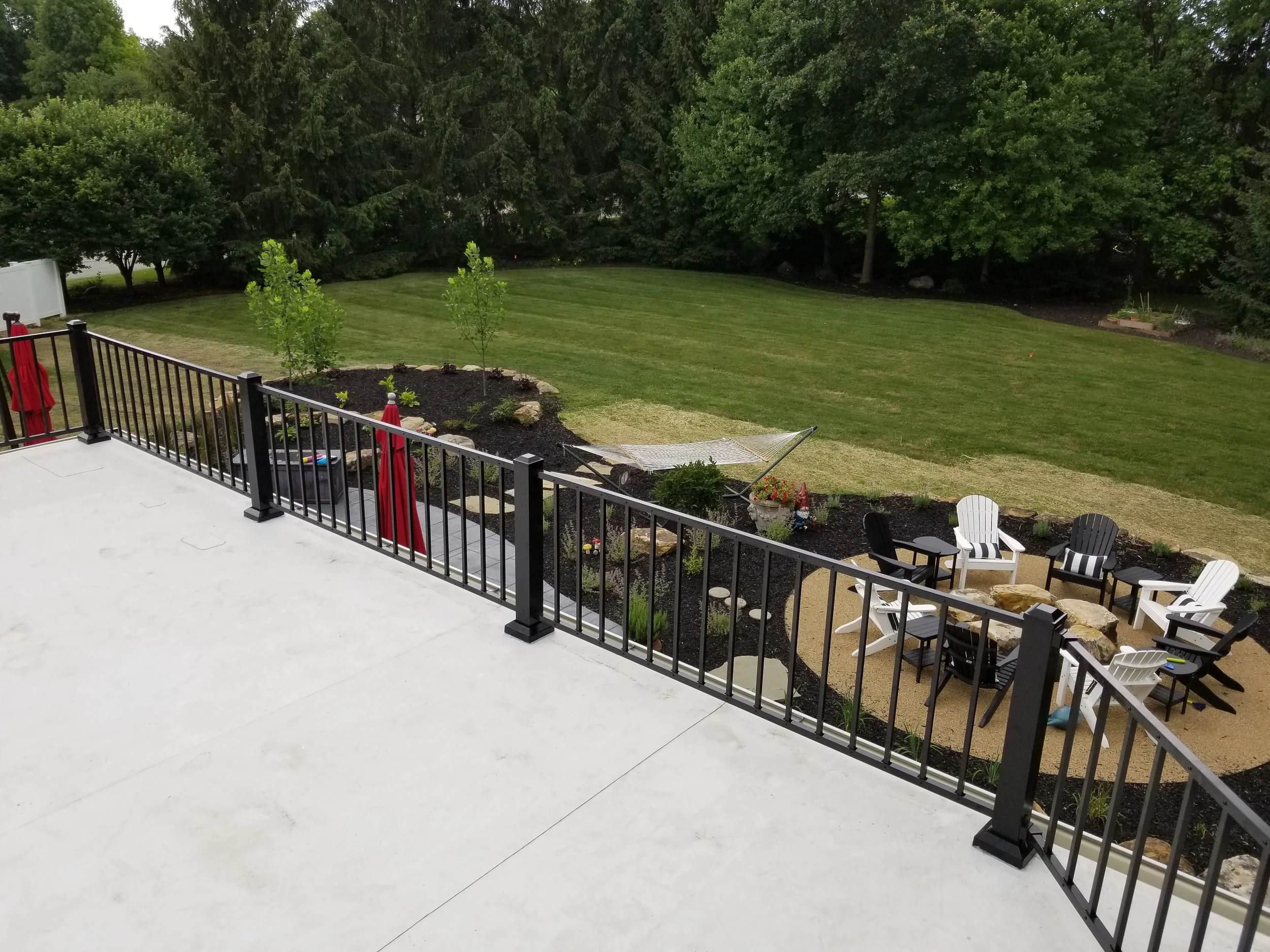 View of a backyard with a deck, a firepit area with chairs, a hammock, a garden bed with plants, and a large grassy lawn bordered by trees.