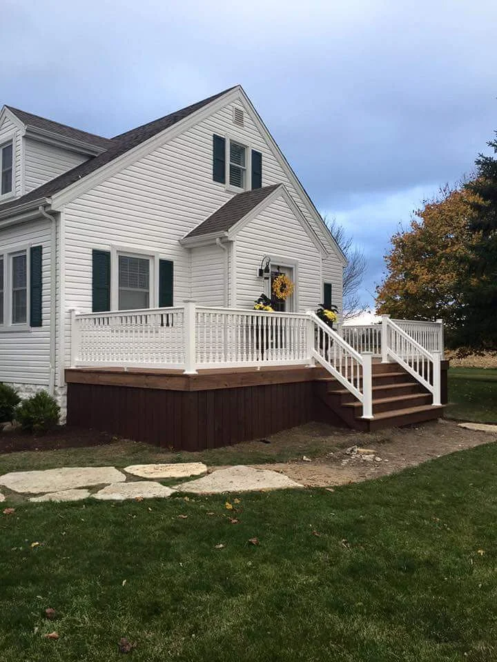 White house with a front porch and stairs, decorated with fall flowers and a wreath, on a well-kept lawn with large stepping stones and a tree with autumn leaves.
