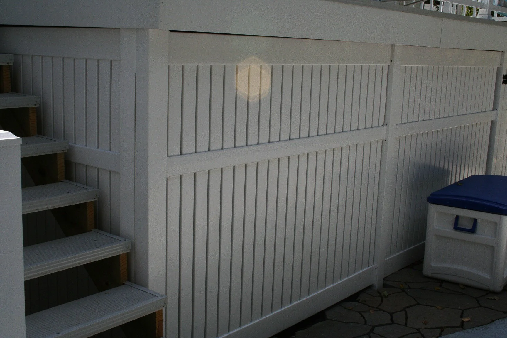 White wooden fence with stairs on the left and a small white storage box with a blue lid on the right. The ground is paved with irregular stones.