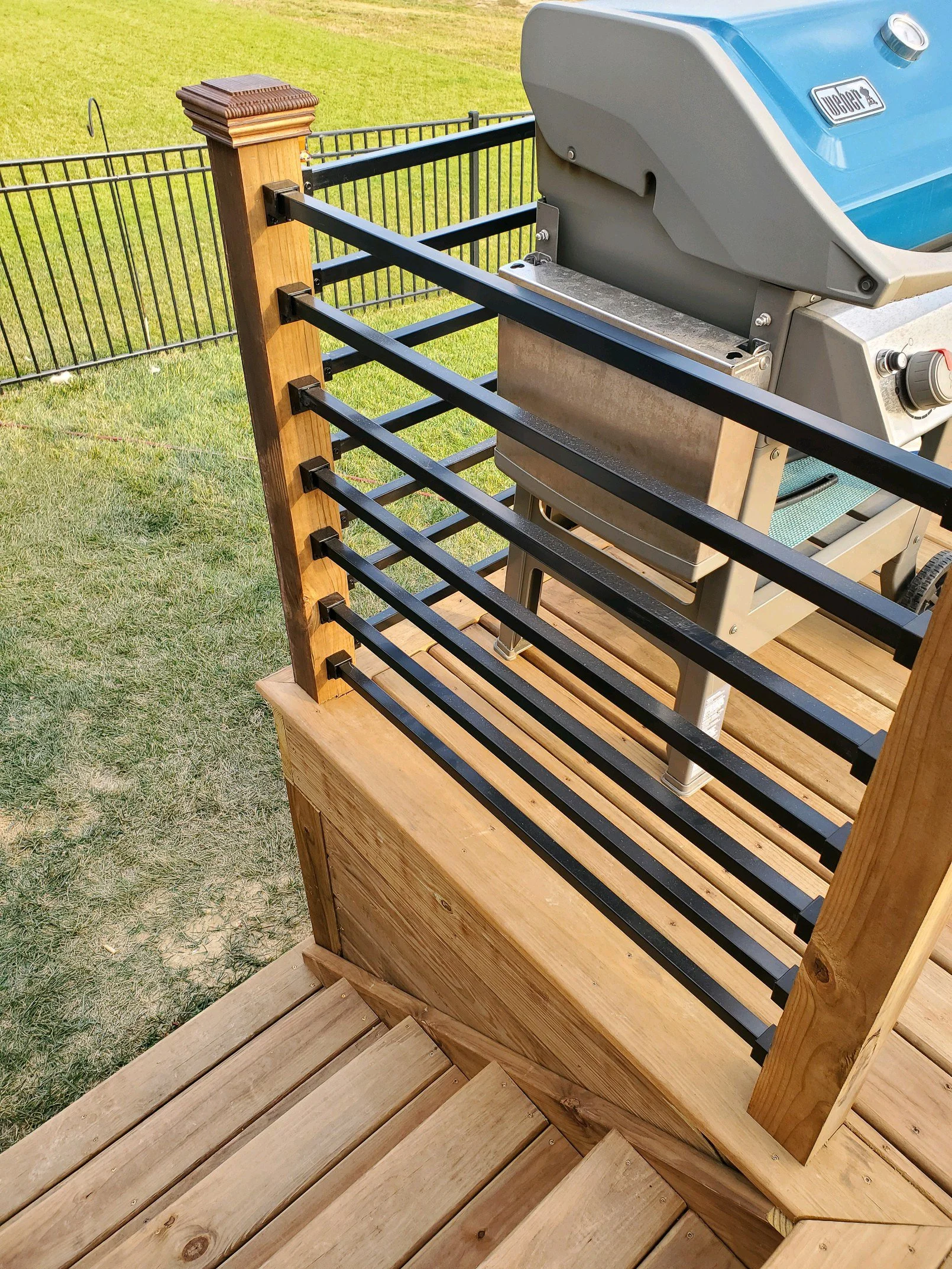 Wooden deck with black metal railing surrounding a gas grill on a grassy yard.