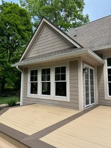 An exterior view of a house with beige siding and four white-framed windows, a sloped roof, and a beige deck in the foreground. Trees are visible in the background.
