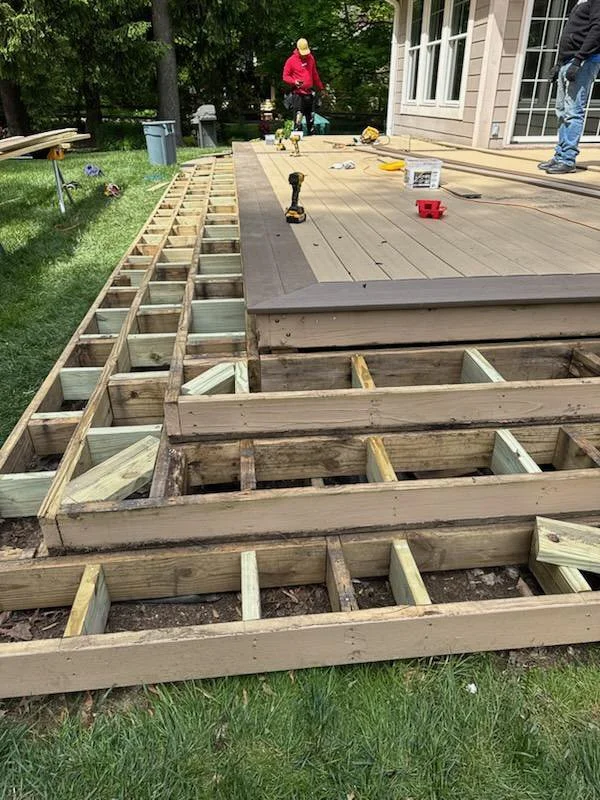 People working on building or repairing a wooden deck, with tools and materials on it, in a backyard with green grass and trees.