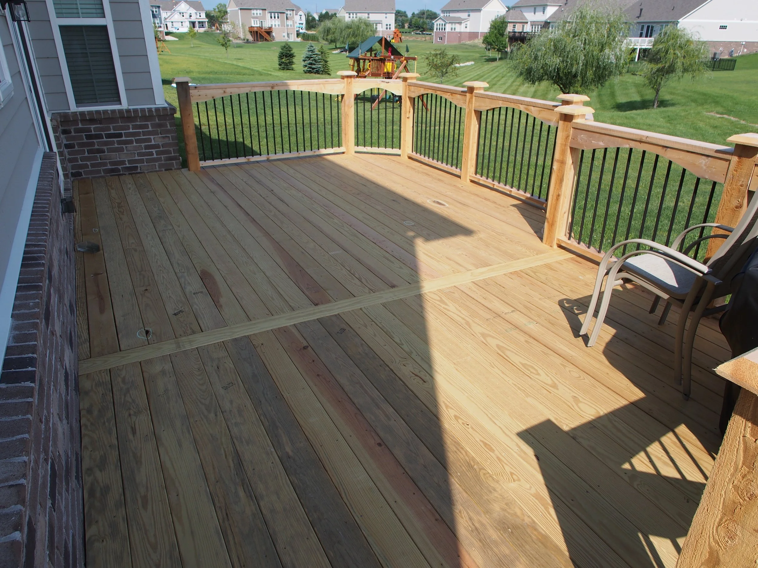 A newly constructed wooden deck with a safety railing overlooking a green backyard with trees and neighboring houses.